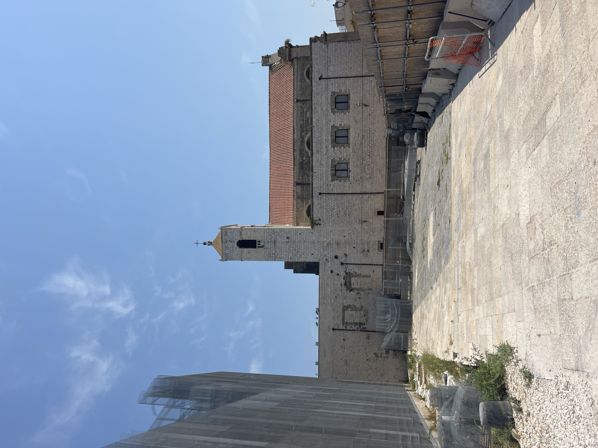 The image depicts an outdoor scene in the city of Bari, Italy, with a church featuring a bell tower and a red tiled roof as the central subject. The building is constructed of light-colored stone blocks. In the foreground, the ground is paved with light-colored tiles, and there is a construction site with scaffolding, netting, concrete barriers, and orange safety fences. The sky is blue with scattered white clouds. The overall impression is of a historic building undergoing renovation or located in an area with ongoing construction.