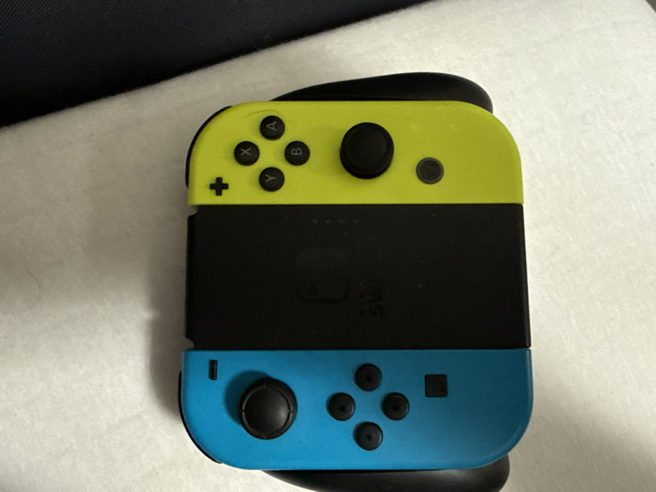 A Nintendo Switch Joy-Con grip is depicted lying flat, viewed from a slightly elevated angle. The left Joy-Con is turquoise blue, featuring a black analog stick, a four-button directional pad, a black minus button, and a black square capture button. The right Joy-Con is neon yellow, displaying a black analog stick, four black face buttons labeled 'X', 'Y', 'A', 'B', a black plus button, and a black circular home button. Both Joy-Cons are attached to a central black grip module. The "NINTENDO SWITCH" logo is visible in white lettering on the black central section. The entire assembly rests on a light-colored, likely white, textured fabric surface. A darker, possibly navy blue or black, fabric element is visible in the upper right background, forming a soft diagonal edge. Shadows are present, indicating an overhead light source. No individuals are present, and no actions or interactions are occurring. The location is Vellmar, Germany.