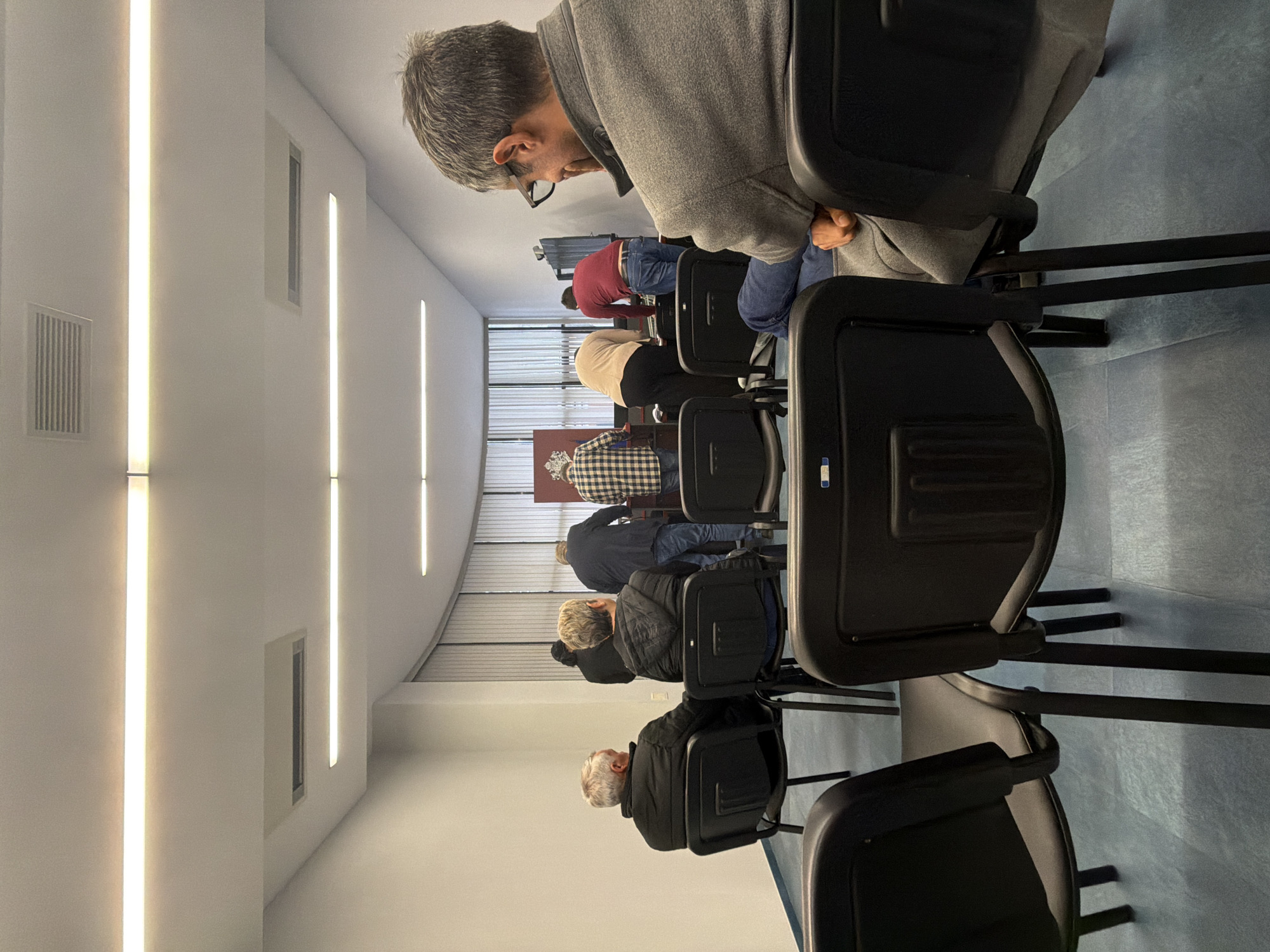 A photograph depicts a row of seated individuals in a room, seemingly an official space or waiting area, located in Plovdiv, Bulgaria. The individuals are seated in black chairs, facing forward. The foreground is dominated by the backs of these chairs, obscuring some of the individuals. The room is brightly lit with long, vertical light fixtures on the left wall. The background features a coat of arms. The floor appears to be covered in blue linoleum. There is a window on the left wall. The room is relatively sterile.