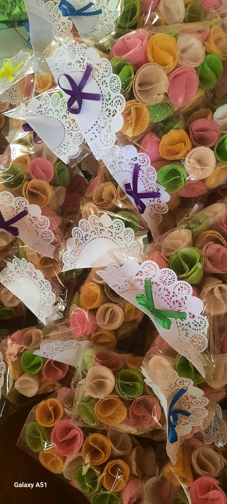 The image displays a close-up, overhead view of numerous individually wrapped, colorful wafer-like treats, resembling small rolled cones or flowers. These treats are in various pastel shades of pink, yellow, green, white, and beige. Each treat is enclosed in clear plastic packaging. Many of the packages are adorned with decorative white paper doilies, often with a small ribbon tied to them in colors such as purple, green, or blue. The treats are densely packed together, filling the frame. The setting appears to be a retail or market environment, likely within a home or small shop in Jatitengah, Indonesia, given the visible text "Galaxy A51" in the lower left corner, which is a common smartphone model used in the region, suggesting a photograph taken by a local. There are no people visible in the image.