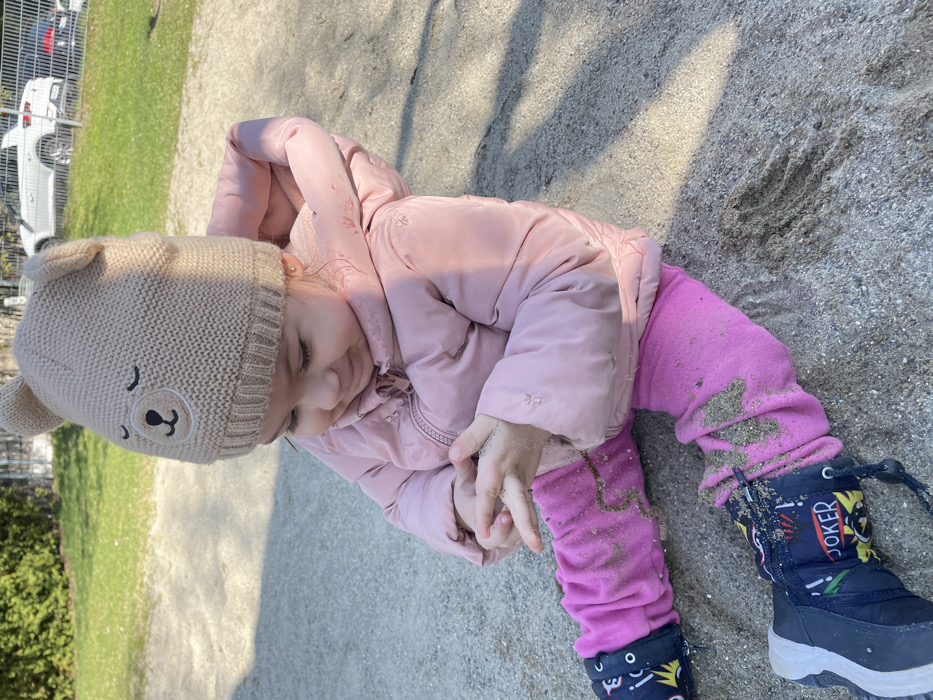 A young child, presumably in Senden, Germany, is seated in a sandpit, observed from a slightly elevated perspective. The child wears a light beige knitted beanie with embroidered sleeping bear facial features and small ears, a light pink padded jacket, fuchsia pants, and dark blue boots decorated with "JOKER" and other graphic motifs. Sand is present on the child's hands, which are clasped together, and scattered across their pants and boots. The child's gaze is directed downwards towards their hands. The immediate environment consists of sand. In the background, green grass and foliage are visible to the left. To the right and further back, a light-colored SUV is parked or situated on a paved surface, with additional vehicles partially visible behind it, bordering what appears to be a public area or park. Distinct shadows indicate direct sunlight.