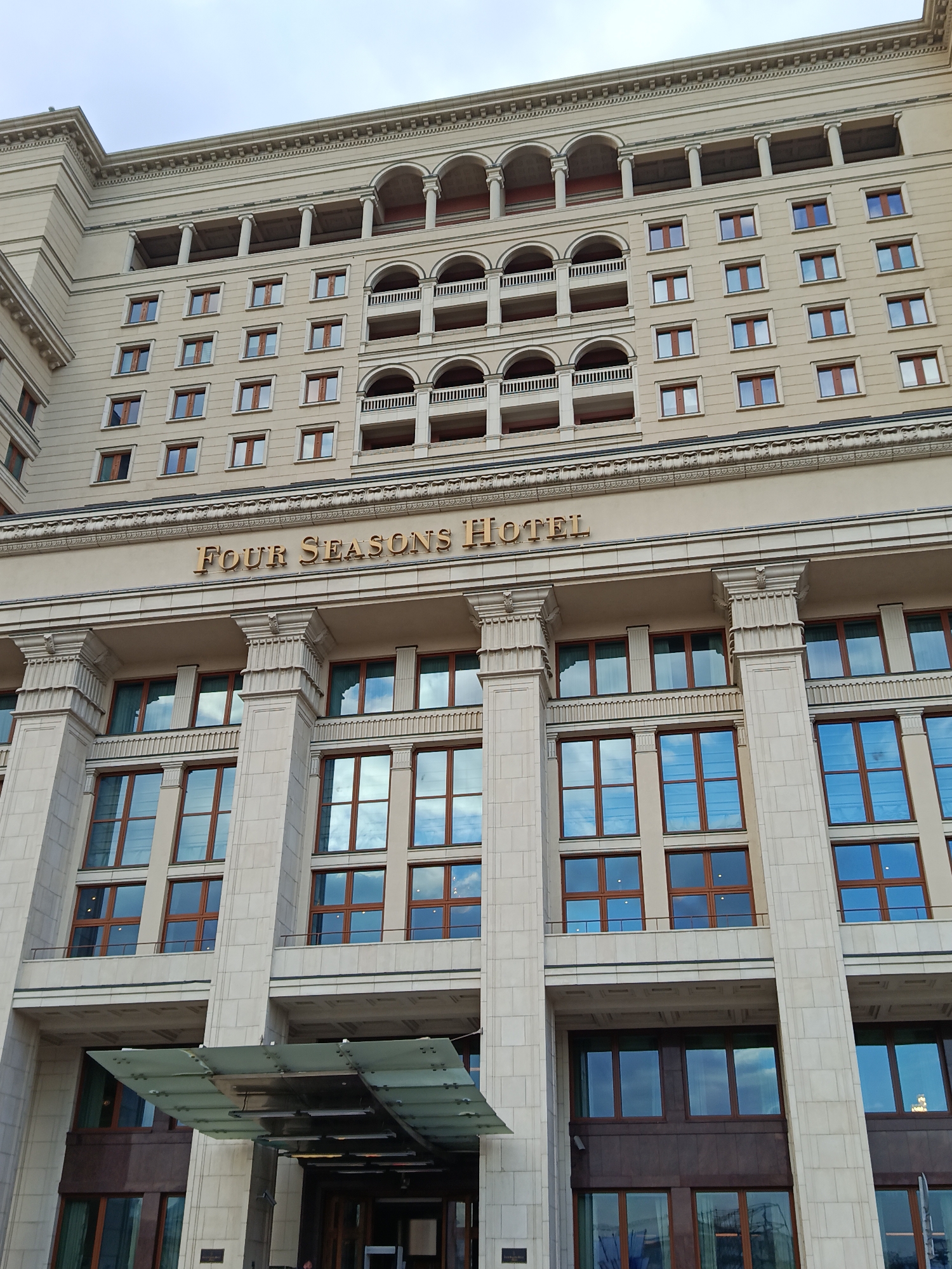 The image shows the exterior of the Four Seasons Hotel in Moscow, Russia. The building is a large, beige-colored structure with multiple stories. It features classical architectural elements such as columns and arched balconies. The facade includes numerous windows reflecting the sky, and the hotel's name is displayed in gold lettering above the entrance. An awning covers the entrance. The sky is overcast.
