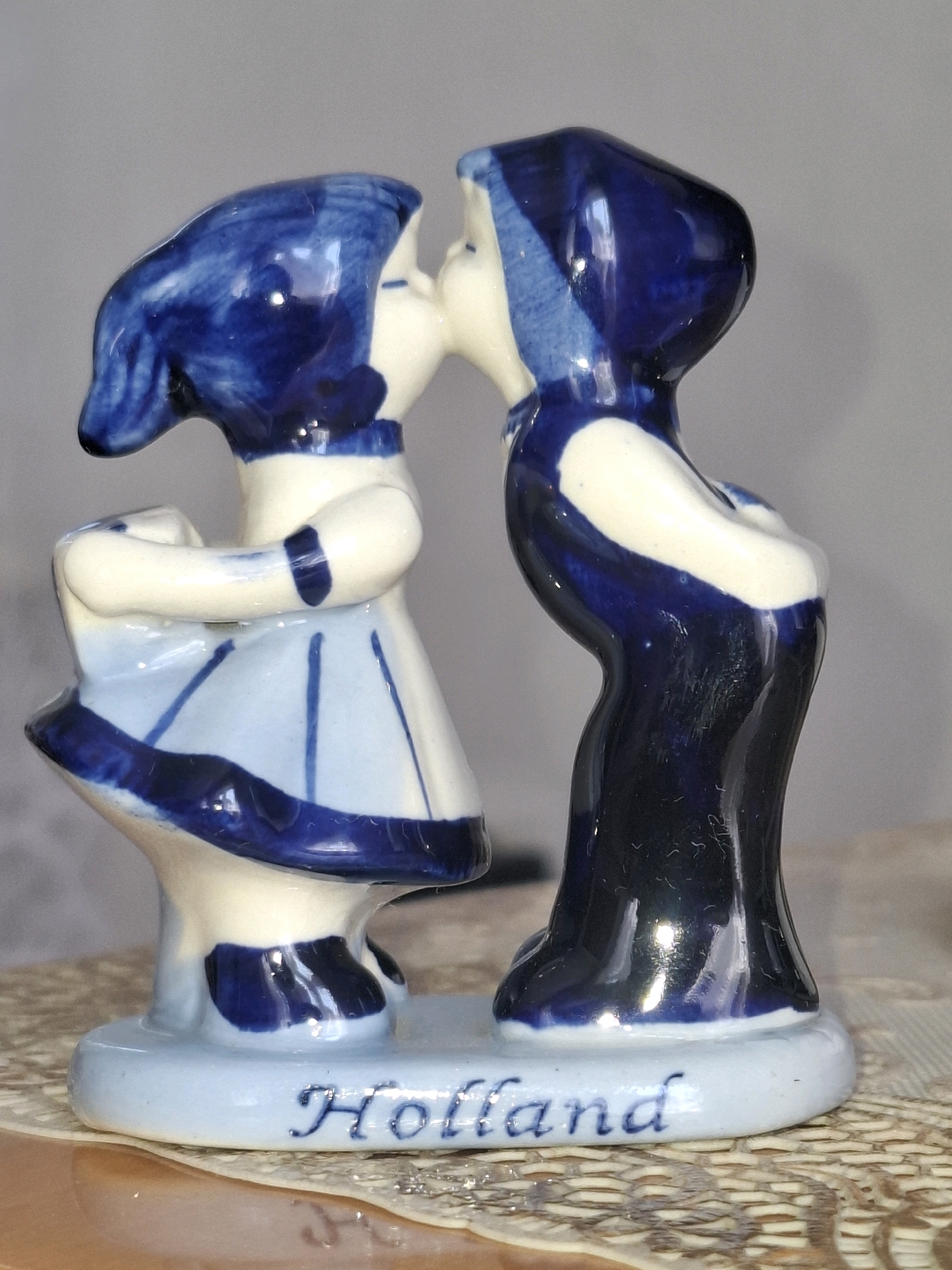 This is a close-up, eye-level shot of a small ceramic figurine depicting a boy and a girl kissing. The figurine is primarily white with blue Delft-style decoration. The girl, on the left, wears a light blue dress with dark blue trim and stripes, and a dark blue bonnet. Her arms are wrapped around her waist. The boy, on the right, wears dark blue trousers and a white shirt with dark blue trim, and a dark blue cap. Both figures have their eyes closed and are leaning towards each other for a kiss. The base of the figurine is light blue and has the word "Holland" inscribed on it in blue script. The background is a neutral gray, and the figurine is placed on a surface with a woven texture, possibly a tablecloth or placemat. The lighting is soft, and there are reflections on the glossy ceramic surface, suggesting artificial light. No specific time of day or weather conditions are discernible. The figurine represents a romantic or innocent moment between two children. The inscription "Holland" indicates a potential origin or theme of the figurine.