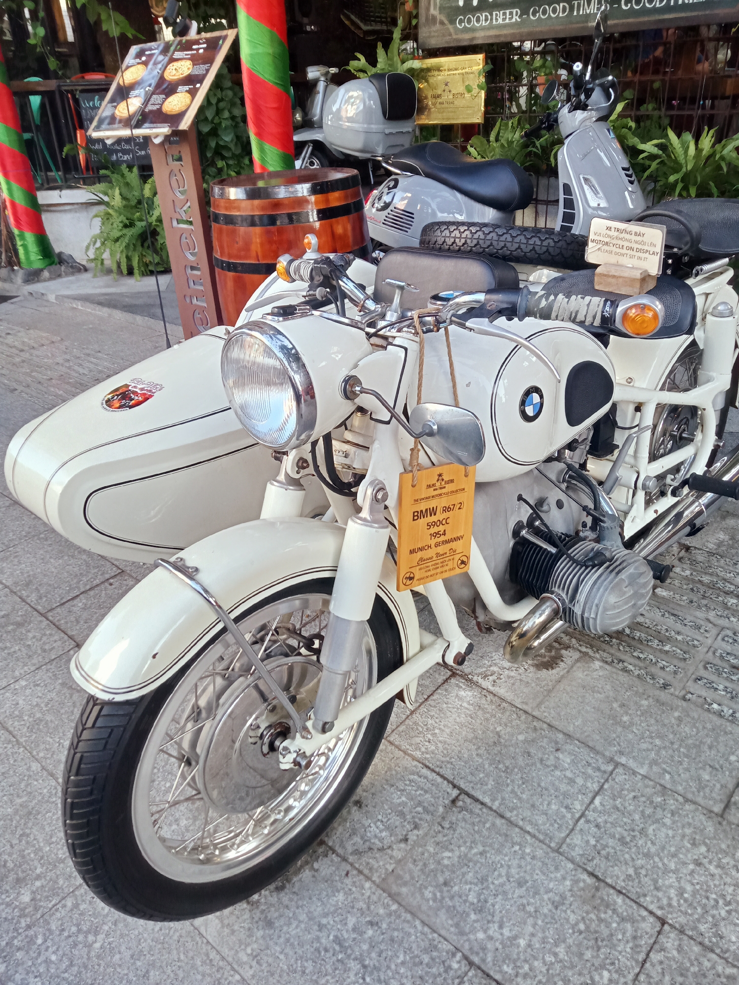 A vintage white BMW motorcycle with a sidecar is parked on a gray tiled surface, likely a sidewalk, in front of a building or establishment. A sign attached to the motorcycle provides information about the vehicle, indicating it is a BMW (R67/2) from 1954, made in Munich, Germany. Another nearby sign reads, "XE TRƯNG BÀY VUI LÒNG KHÔNG NGÔI LÊN MOTORCYCLE ON DISPLAY PLEASE DON'T SIT IN IT”. To the left of the motorcycle is a Heineken sign with pizza ads. In the background, two grey scooters are parked, with signage indicating the location is Nha Trang, Vietnam (PALAIS BISTRO NHA TRANG). In the top right, a sign reads “GOOD BEER - GOOD TIMES”. There are red and green poles and ferns around.