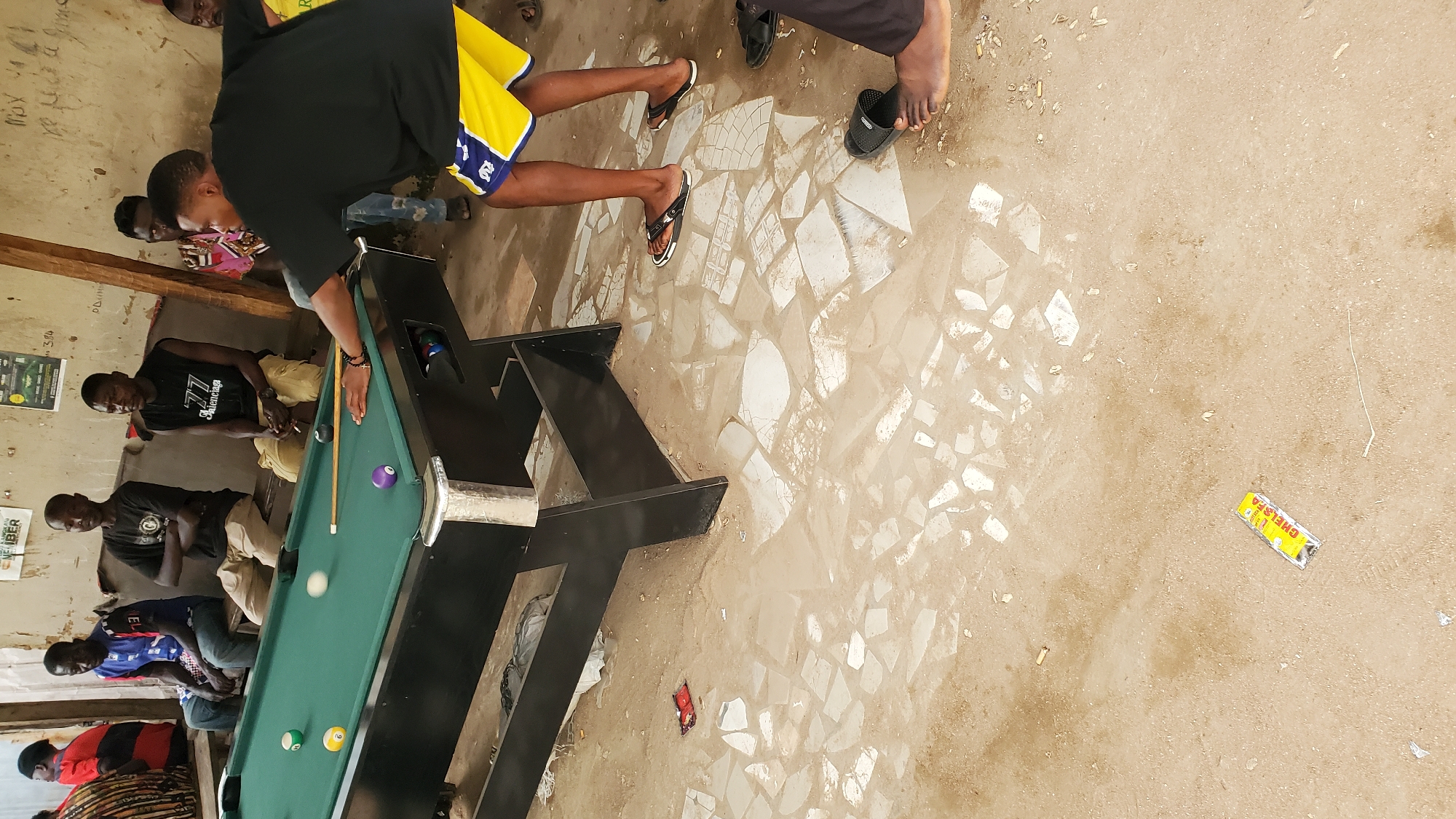 In Jalingo, Nigeria, a group of men are gathered around a pool table in an outdoor, semi-covered area with a dirt and broken tile floor. The scene appears to be informal, possibly a local hangout or recreational spot. The men are dressed casually, with some wearing t-shirts and shorts, others jeans. One man is actively playing pool, leaning over the table and preparing to strike a ball with a cue. Other men are observing the game, seated on a bench behind the pool table or standing nearby. The pool table is green, with several colored balls on it, including a white cue ball that appears to be in motion. In the background, there are walls with posters and graffiti, suggesting a well-worn or frequented location. The overall atmosphere is relaxed and social, with the game of pool being the central activity.