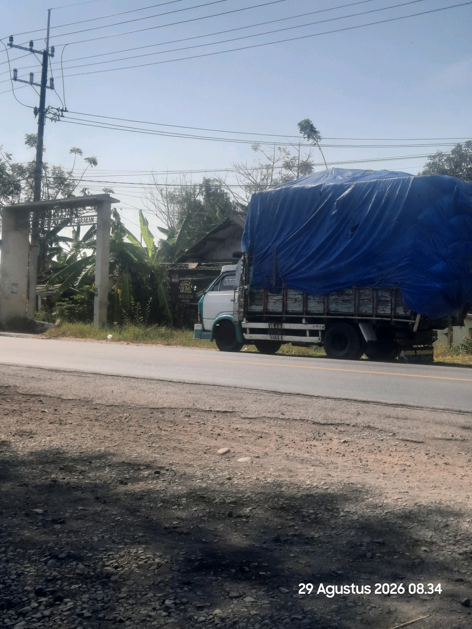 A truck covered with a blue tarp is parked on the side of a road in Sukokerto, Indonesia. The truck is white with a teal trim on the lower part of the cab. The truck bed is wooden with visible signs of wear and tear. The truck has dual rear wheels and appears to be a medium-duty vehicle. In the background, there is a concrete archway with text that is partially obscured by vegetation. The archway leads to a property with houses and lush green banana trees. A utility pole with multiple wires runs along the top left of the image. The road is paved and has yellow lane markings. The ground to the left of the road is covered with gravel and shadows. The sky is clear and blue. The date stamp "29 Agustus 2026 08.34" is visible in the bottom right corner.