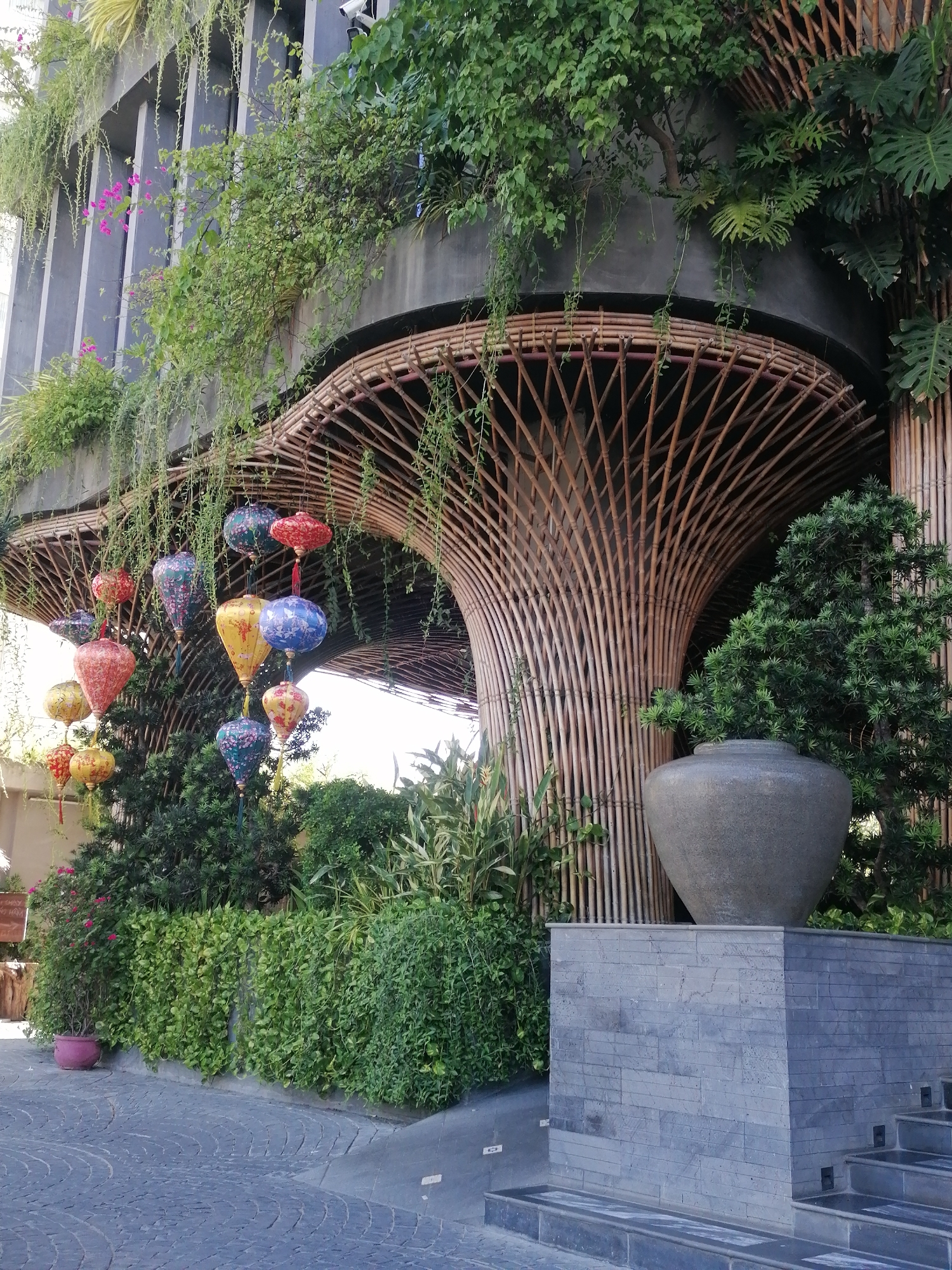 The scene captures a lush, architectural detail in Nha Trang Ward, Vietnam. The focal point is an organically shaped structure composed of thin, interwoven bamboo poles that form an archway. From this structure hang several colorful lanterns, adding a vibrant touch to the setting.
The architecture is integrated with abundant greenery; plants cascade down from the building above, and shrubs and potted flowers adorn the base of the structure. A large, gray urn sits atop a stone block wall, nestled among the foliage. The building itself has a modern design, with tall, vertical elements.
The foreground consists of a paved path that leads up to the structure, suggesting a public space or entrance to a building. The overall impression is one of a harmonious blend of natural and man-made elements.