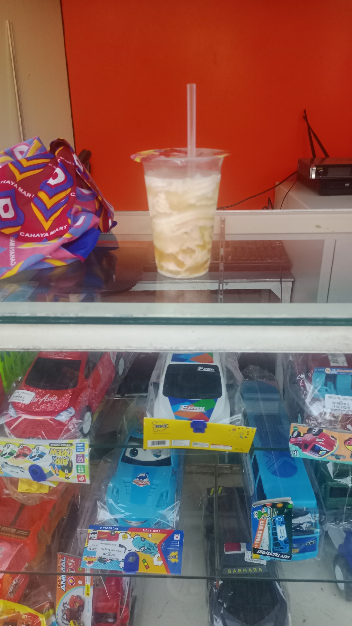 This image shows a display case filled with various toys, predominantly toy cars and buses, inside a store. The toys are mostly packaged in clear plastic, and some are brightly colored. Above the toys, a clear plastic cup with a straw contains a creamy, pale yellow beverage, possibly a dessert drink, with some solid pieces visible within. To the left of the drink, a folded cloth bag with a geometric pattern in red, yellow, blue, and white is visible. In the background, a red wall serves as a backdrop, and on a shelf behind the drink, a black electronic device, possibly a DVD player or similar, is present. The overall impression is of a retail setting where toys and refreshments are sold. The specific location of Pakemitan, Indonesia, is not directly indicated by any visual cues within the image itself.