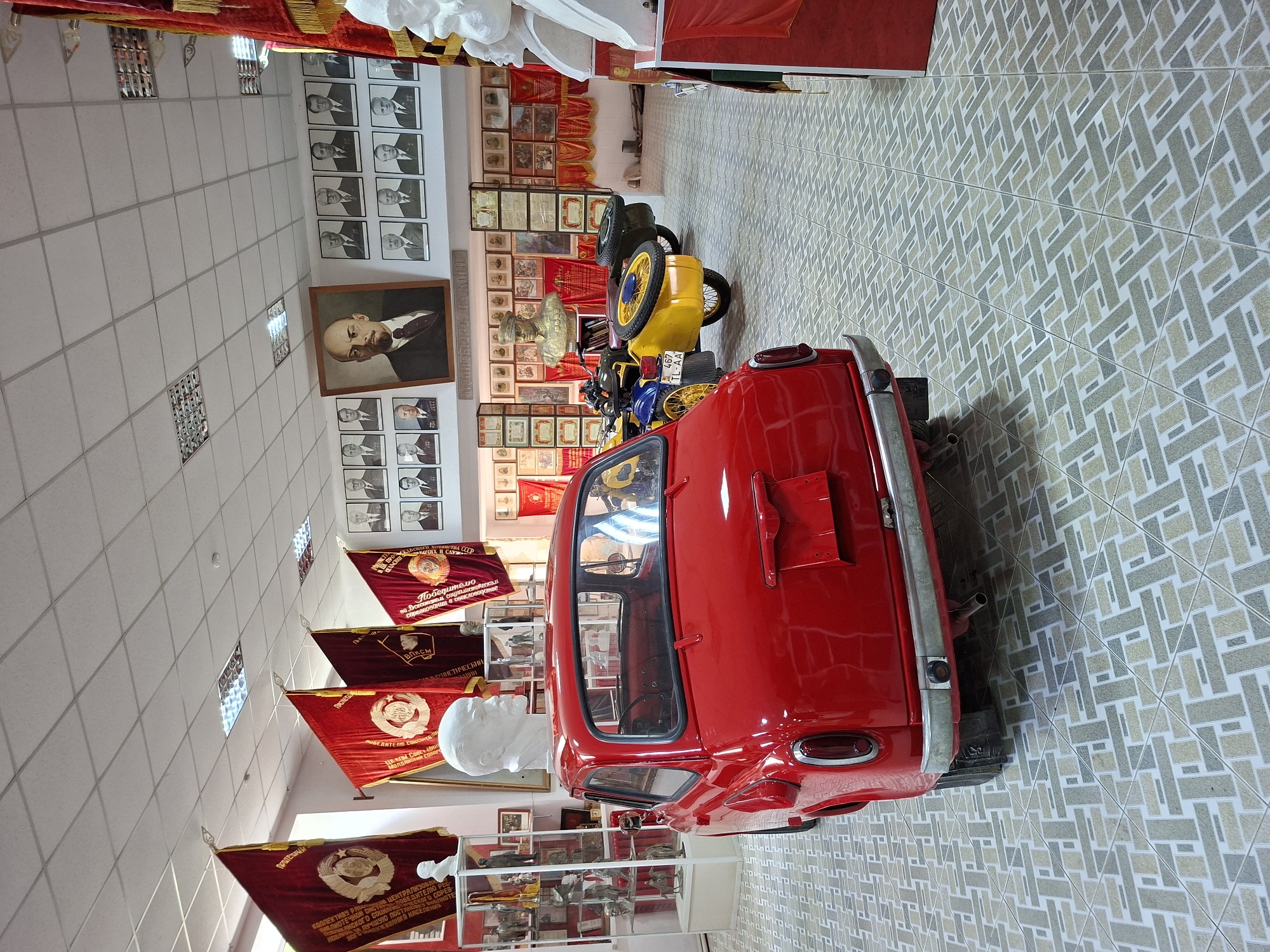 The image depicts the interior of a museum, likely a local history or political museum, in Ialoveni, Moldova. The scene is dominated by a bright red, vintage car parked centrally in the foreground. To the left of the car, a glass display case contains various artifacts. Behind the car, a yellow motorcycle with a sidecar is visible. The walls are adorned with framed portraits of historical figures, predominantly of Soviet leaders like Lenin, along with numerous flags and banners, some featuring Soviet symbols. Busts of notable figures are also displayed. The floor is tiled in a geometric pattern. The overall atmosphere suggests a preserved historical exhibit.