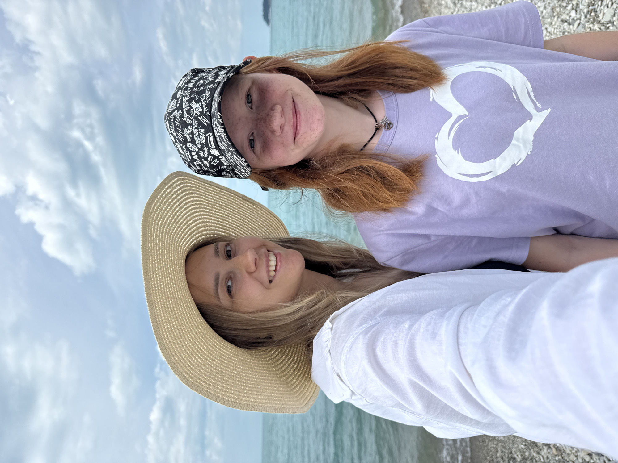 Two young women, likely friends or sisters, pose for a selfie at the beach in Nebug, Russia. The woman on the left wears a wide-brimmed straw hat and a white linen shirt, her blonde hair framing her smiling face. The woman on the right has striking red hair, freckles, and wears a black baseball cap with a white graphic pattern. She is wearing a light purple t-shirt with a large white heart design. They stand on a pebbled beach with the calm, blue-green waters of the Black Sea behind them, under a cloudy sky. The overall atmosphere is relaxed and summery.