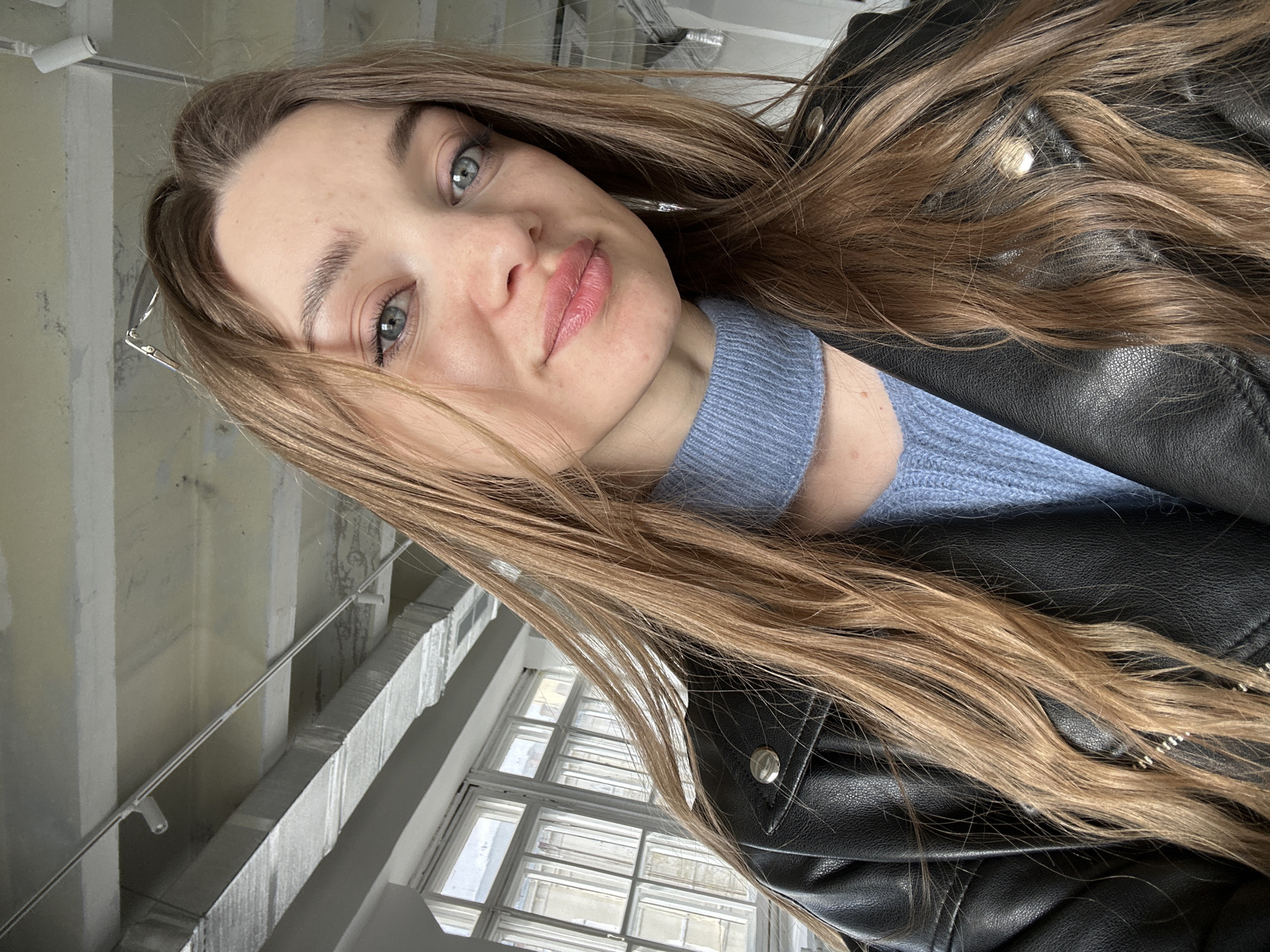A young woman is photographed indoors. She has long, light brown hair, blue eyes, and fair skin. She wears a light blue sweater and a black leather jacket. The background shows a white, industrial-style ceiling with exposed ductwork and track lighting. Natural light enters through a grid-patterned window. The location in the city of Kazan, Russia, is not discernible from the image.