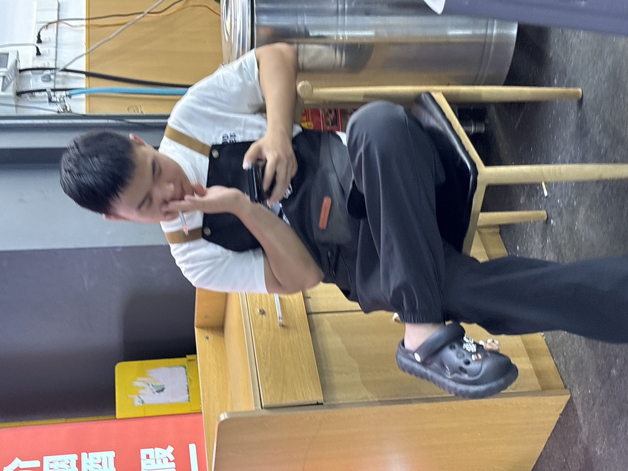 A man is seated on a chair indoors. He is wearing a white t-shirt, a black apron, and black pants, with black Croc-style shoes. He is holding a black object, possibly a phone, in his right hand, and his left hand is near his face. The man is seated next to a light brown wooden cabinet. The location appears to be in the Yuzhong District of China, based on the visible Chinese characters on the red sign. A metal container is positioned behind the chair.
