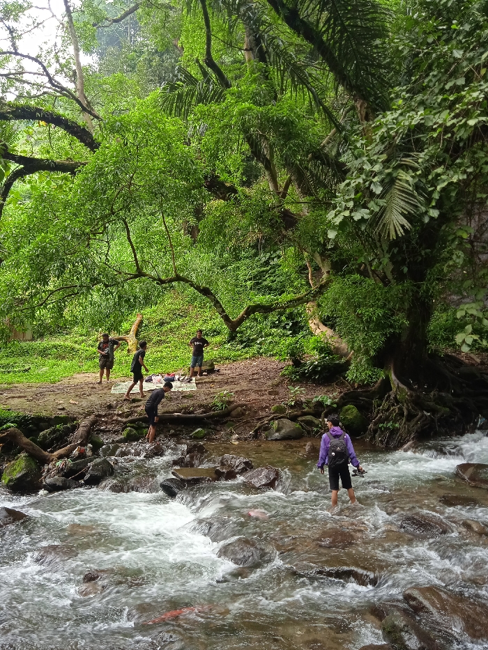 The image shows a group of people standing and walking in a shallow river in Tawangmangu, Indonesia. The river is rocky and has rapids flowing over the rocks. The people are standing on the rocks, and some are walking across the river. There is dense green vegetation and trees on both sides of the river. The coordinates -7.654, 111.136 are approximately in the area of Tawangmangu, Indonesia. One person is wearing a purple jacket, a black backpack, and black shorts. Another person is wearing a blue shirt and black shorts. Another person is wearing a red shirt and black shorts. Another person is wearing a black shirt and black shorts. Another person is wearing a black shirt and black shorts. Another person is wearing a white shirt and black shorts. Another person is wearing a white shirt and black shorts. There is a white blanket and some belongings lying on the ground next to the river. The image was taken from a vantage point overlooking the river.