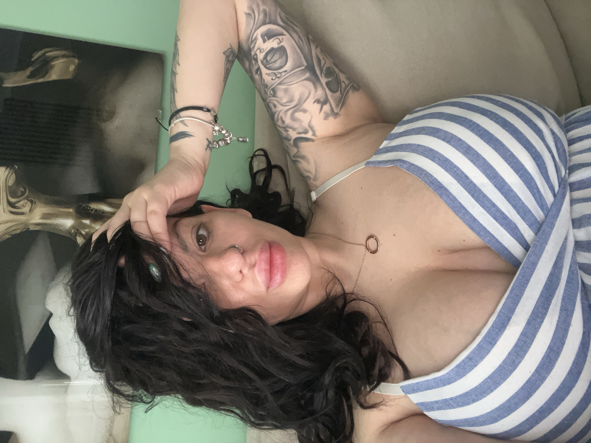 A fair-skinned person is lying down, viewed from an overhead perspective. The person has dark, wavy hair and dark eyes. They are wearing a blue and white striped garment with thin straps and a necklace with a circular pendant. Their left arm, featuring tattoos, is raised with the hand resting on their head. Bracelets adorn the wrist. The background includes a light green wall and a piece of art with monochrome imagery. The context does not suggest a location in Plovdiv, Bulgaria.