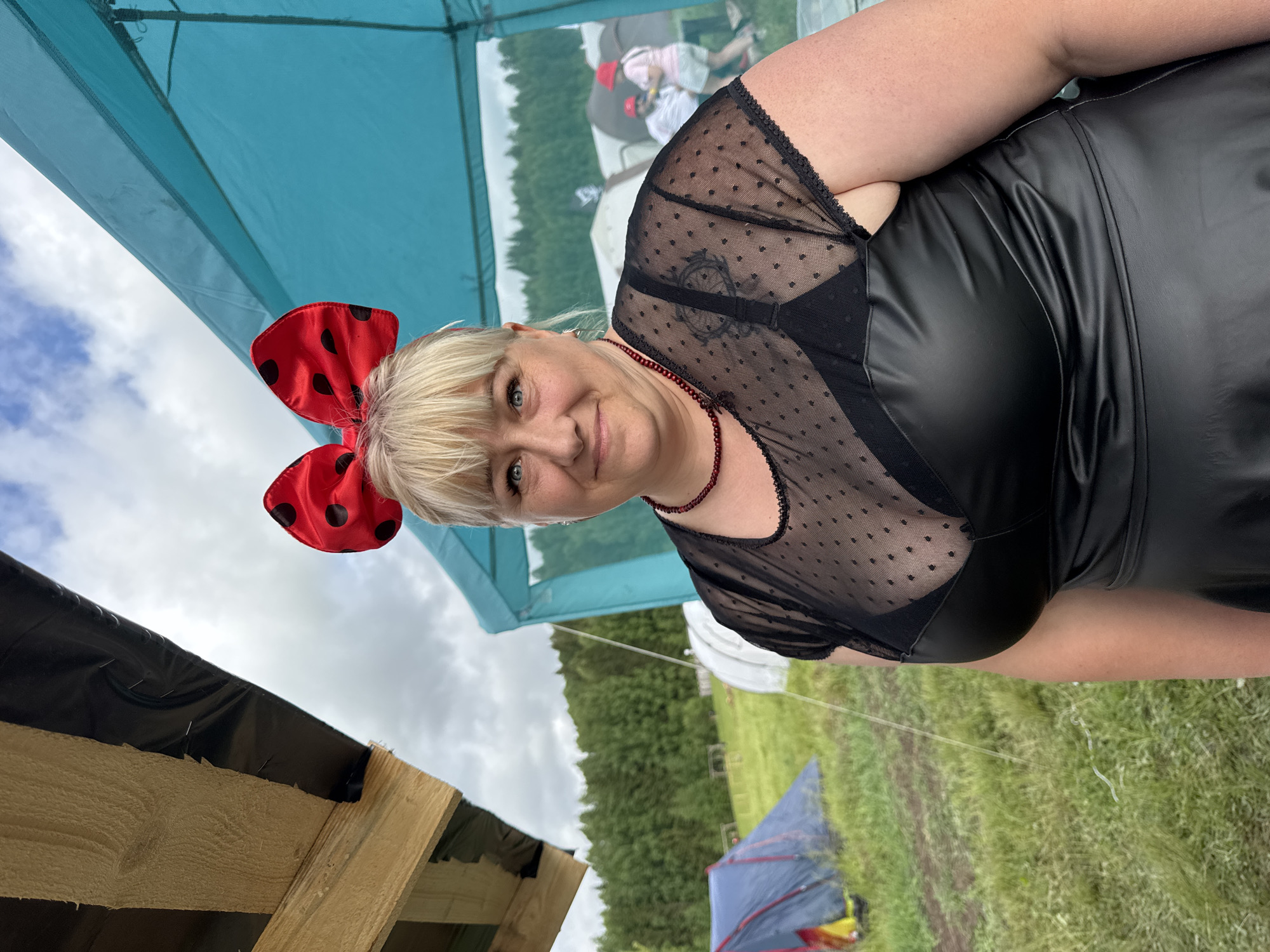 A woman with blonde hair, light-colored eyes, and a fair complexion looks directly at the camera with a slight smile. She wears a large red bow with black polka dots on her head, resembling Minnie Mouse ears. Her attire consists of a black sheer top with small dots, a black bra visible underneath, and a black faux leather or pleather dress. A dark red beaded necklace is around her neck, and a circular tattoo is visible on her upper arm through the sheer fabric.
The setting is outdoors, likely a festival, camping event, or market, under a partly cloudy sky during the daytime. A light blue canopy is prominent in the upper left corner of the image. The background features green trees and grassy ground, with a blue and yellow tent and another light-colored structure visible in the distance. A blurred image of a person in a red hat can be seen behind the subject. In the foreground, wooden planks covered with black material are present, possibly part of a makeshift stand or shelter.
The location context for this scene is Большая Кивара, Russia. There is no visible text in the image.