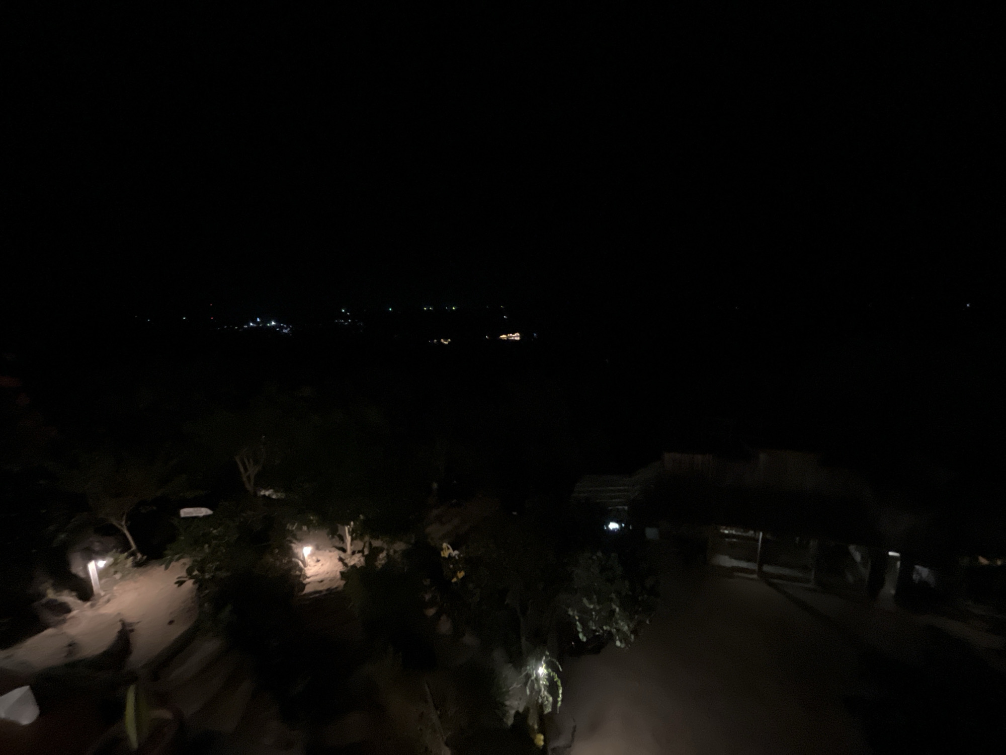 The image captures a dimly lit outdoor scene at night, likely in a rural or resort area, possibly in Karimunjawa, Indonesia, given the context. The foreground features a pathway illuminated by a few artificial lights. To the left, a pathway curves, bordered by dark foliage. On the right side of the pathway, there is a structure with a roof, possibly an open-air pavilion or building, with some lights visible within its interior. In the middle ground, more trees and vegetation are present. In the far distance, a faint line of lights suggests a settlement or a town. The overall impression is one of a quiet, nocturnal environment.