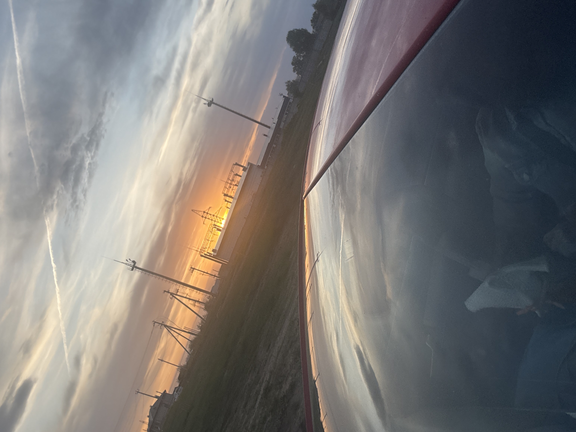 The image shows a sunset view from what appears to be the inside of a vehicle, possibly a car. The vehicle's dashboard or exterior surface is visible in the foreground. The sky is filled with clouds illuminated by the setting sun, creating an orange and yellow glow. In the background, there are electrical power lines and towers. The ground appears to be grassy or unpaved. The location is likely in Chaikovsky, Russia, given the presence of electrical infrastructure common in industrial areas, and the general landscape of the surrounding area. The reflection on the vehicle's surface suggests that there might be a person in the vehicle, but the details are unclear.