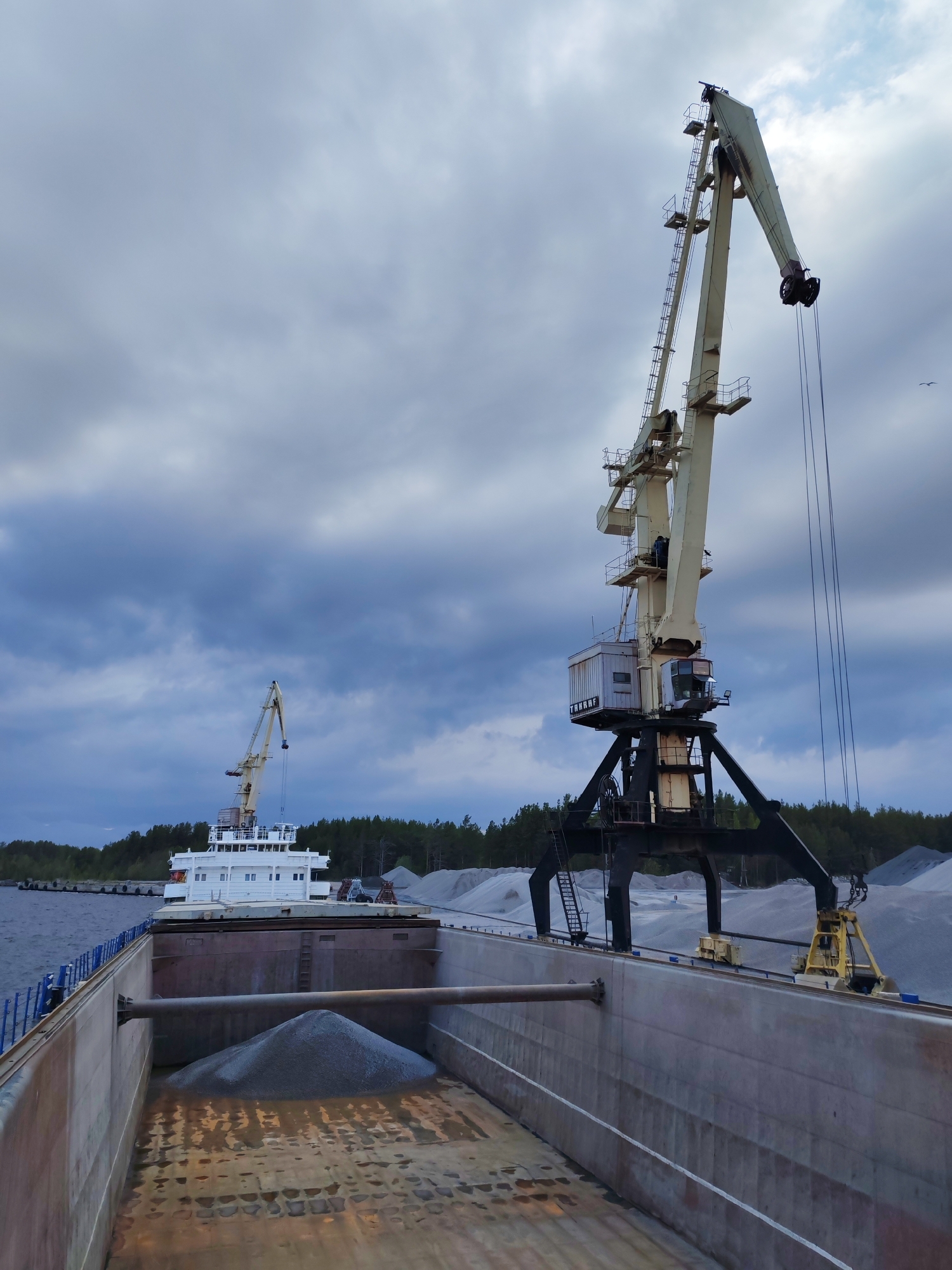 The image shows a cargo ship being loaded with gravel in Shal'skii, Russia. Two large, off-white cranes are positioned alongside the ship. The ship's cargo hold is visible, with a small mound of gravel already loaded. In the background, a stockpile of gravel and a line of trees are visible under a cloudy sky. The ship is docked in the water, with a glimpse of the shoreline and another smaller ship in the distance.