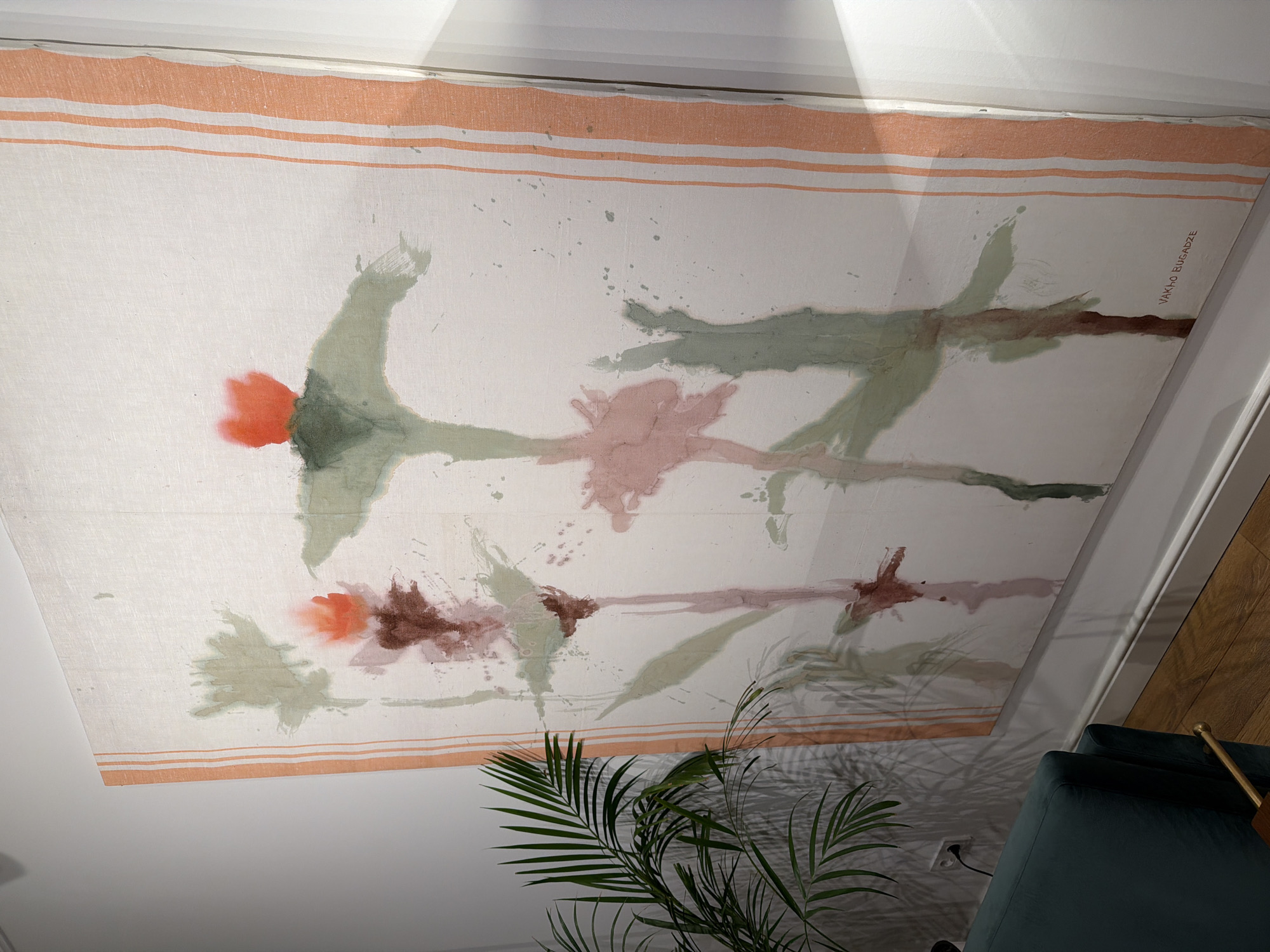The image shows a room interior featuring a large, rectangular art piece on what appears to be a ceiling or a wall. The artwork has a white base with peach-colored stripes along the edges. The art depicts abstract floral or plant-like forms in red, brown, pink, and green hues with splatter effects. The artwork has the artist's name "JAKHO BUGADZE" inscribed in the corner. In the foreground, part of a houseplant with green fronds is visible. Also visible is a portion of a green armchair or sofa and a golden-colored object, possibly part of the furniture. The environment suggests an indoor setting, possibly a residence or an art gallery, in Tbilisi, Georgia, given the artist's name and location.