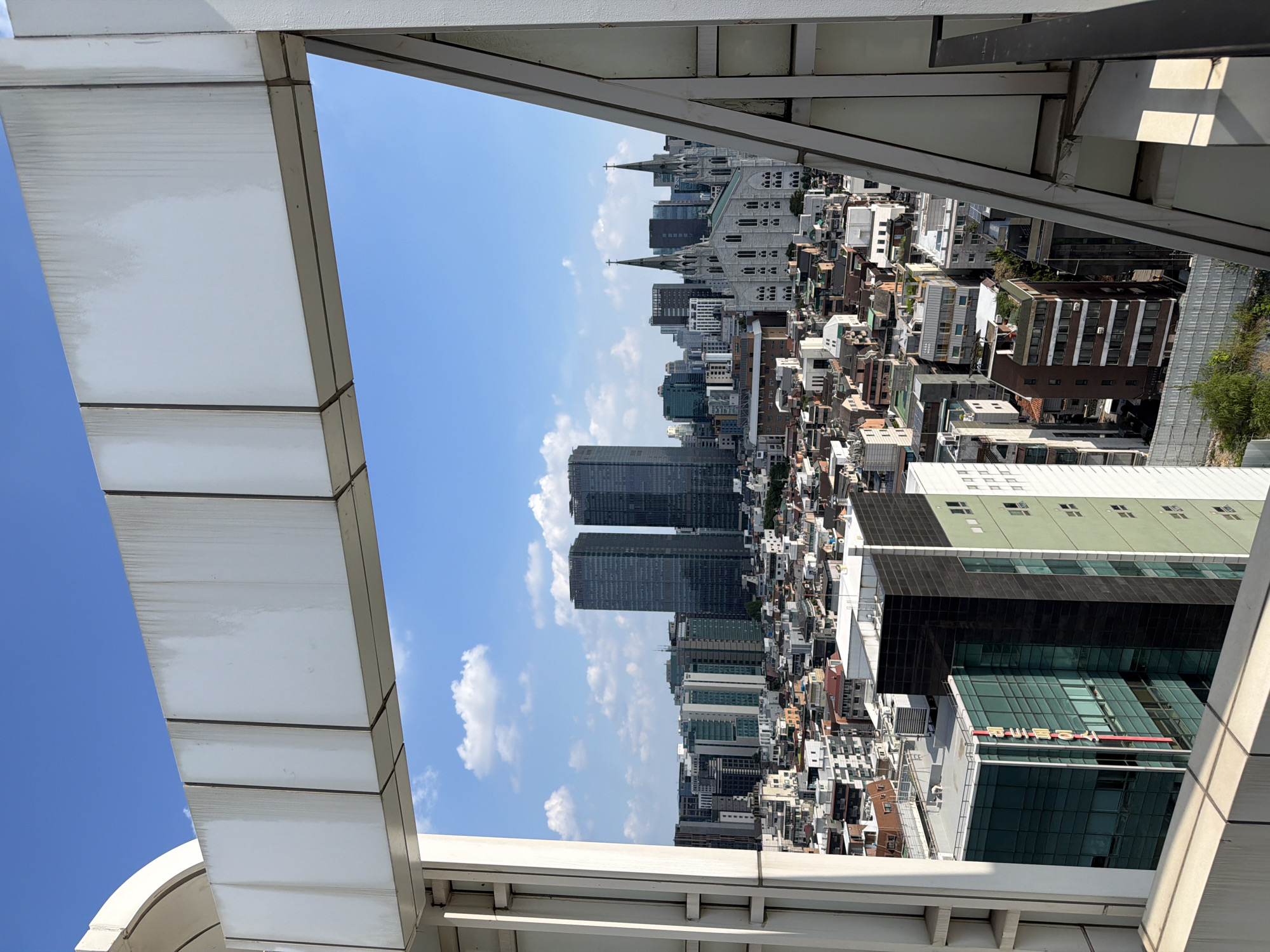The image is a medium-angle, outdoor, daylight shot of the Seoul skyline, viewed from a slightly elevated perspective, possibly a rooftop or a balcony. The foreground is dominated by a white, architectural structure that frames the view of the city. This structure has visible signs of wear, with streaks of dirt and discoloration on its panels, particularly on the upper horizontal beam.
The cityscape behind the foreground structure reveals a dense urban environment with a variety of buildings. Two tall, dark, rectangular skyscrapers stand prominently in the middle ground, side-by-side. To the right of these, a large, white, Gothic-style cathedral with twin spires is a significant focal point. Surrounding these larger structures is a complex patchwork of smaller buildings, many with pitched roofs, suggesting a mix of older and newer developments.
In the middle left of the frame, a modern, multi-story building with a green glass facade is visible. One of its vertical sides has red Korean lettering, possibly a company name or sign. Other buildings in the scene vary in height and architectural style, with some appearing to be residential and others commercial. The sky is a clear blue with scattered white cumulus clouds.
No people are directly visible in the image. The scene is static, capturing a moment in time of the urban landscape. The overall impression is one of a bustling, densely populated city with a mix of architectural influences. The specific location within Seoul can be inferred to be an area with a mix of modern high-rises, older residential buildings, and significant religious architecture, suggesting a central or historically significant district. The presence of the Gothic cathedral and the distinct skyscrapers could potentially pinpoint the location further for someone familiar with Seoul's landmarks.