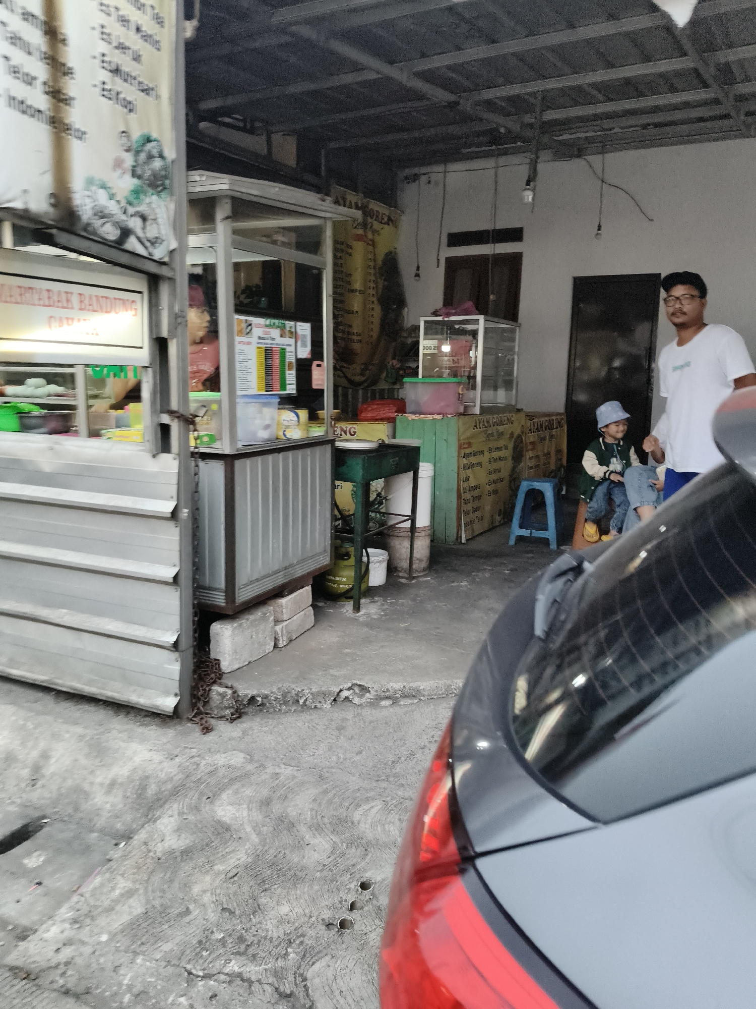 The image depicts a street-side food stall in South Tangerang, Indonesia. A man wearing glasses and a white t-shirt stands to the right of the frame, looking towards the left. Beside him, a young child in a blue hat and jacket sits on a small blue stool, facing the viewer and smiling. The stall itself is a metal structure with glass display cases and hanging menus listing various food and drink items. Behind the stall, a wall with windows and doors is visible, along with exposed electrical wiring and light fixtures on the ceiling. The ground in front of the stall is concrete with a patterned texture. A portion of a dark-colored car is visible in the foreground, with its taillight on the right side of the frame. The overall impression is that of a bustling local eatery.