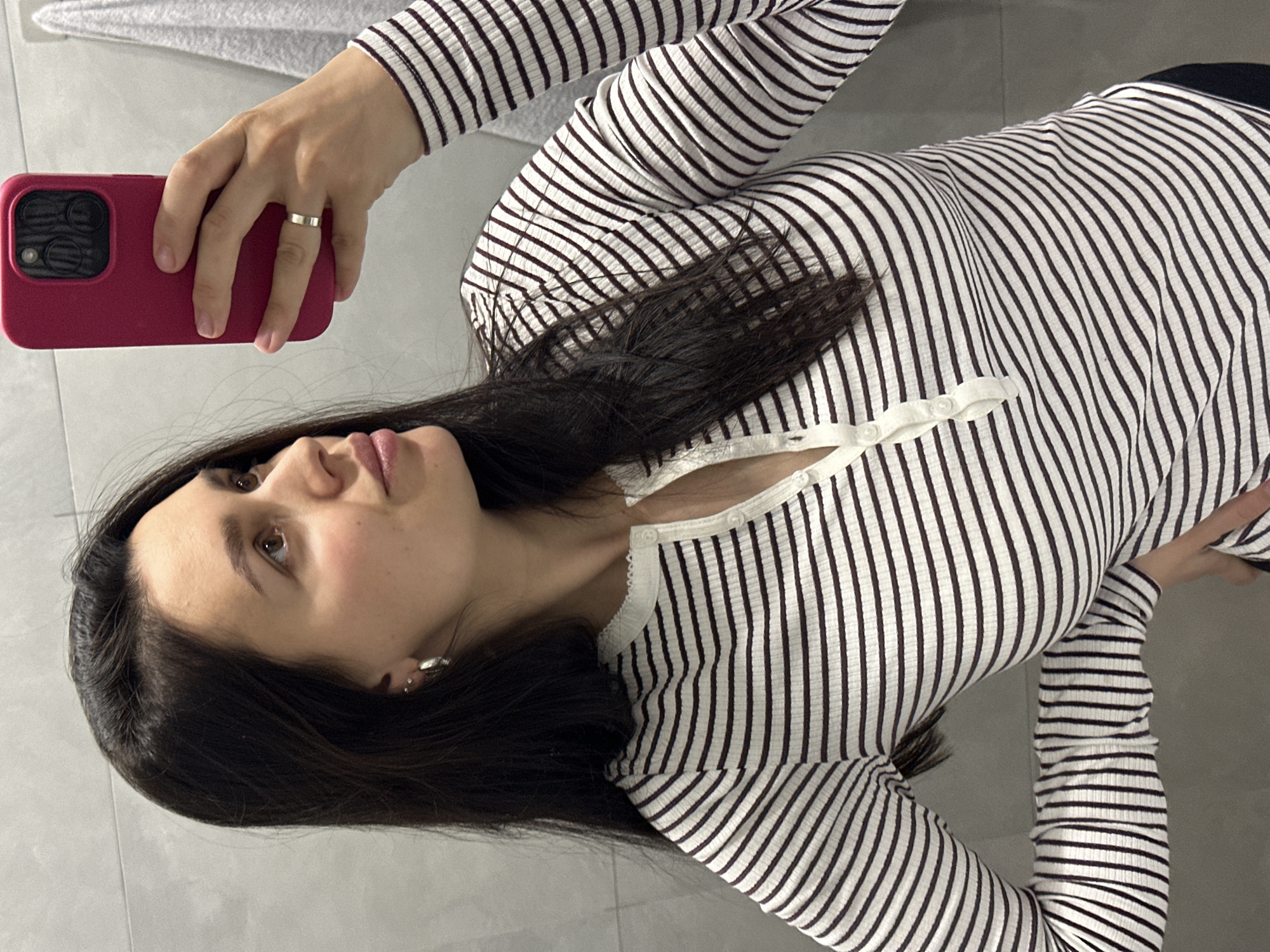 A young woman is taking a selfie in a location with grey tile flooring. She is holding a red phone with a large camera bump in her left hand. She is wearing a long-sleeved, horizontally striped white and brown top with white buttons at the neckline. She has dark hair, light skin, and is wearing a silver ring on her left ring finger. The image provides no information to confirm or deny that the photo was taken in Chaikovsky, Russia.