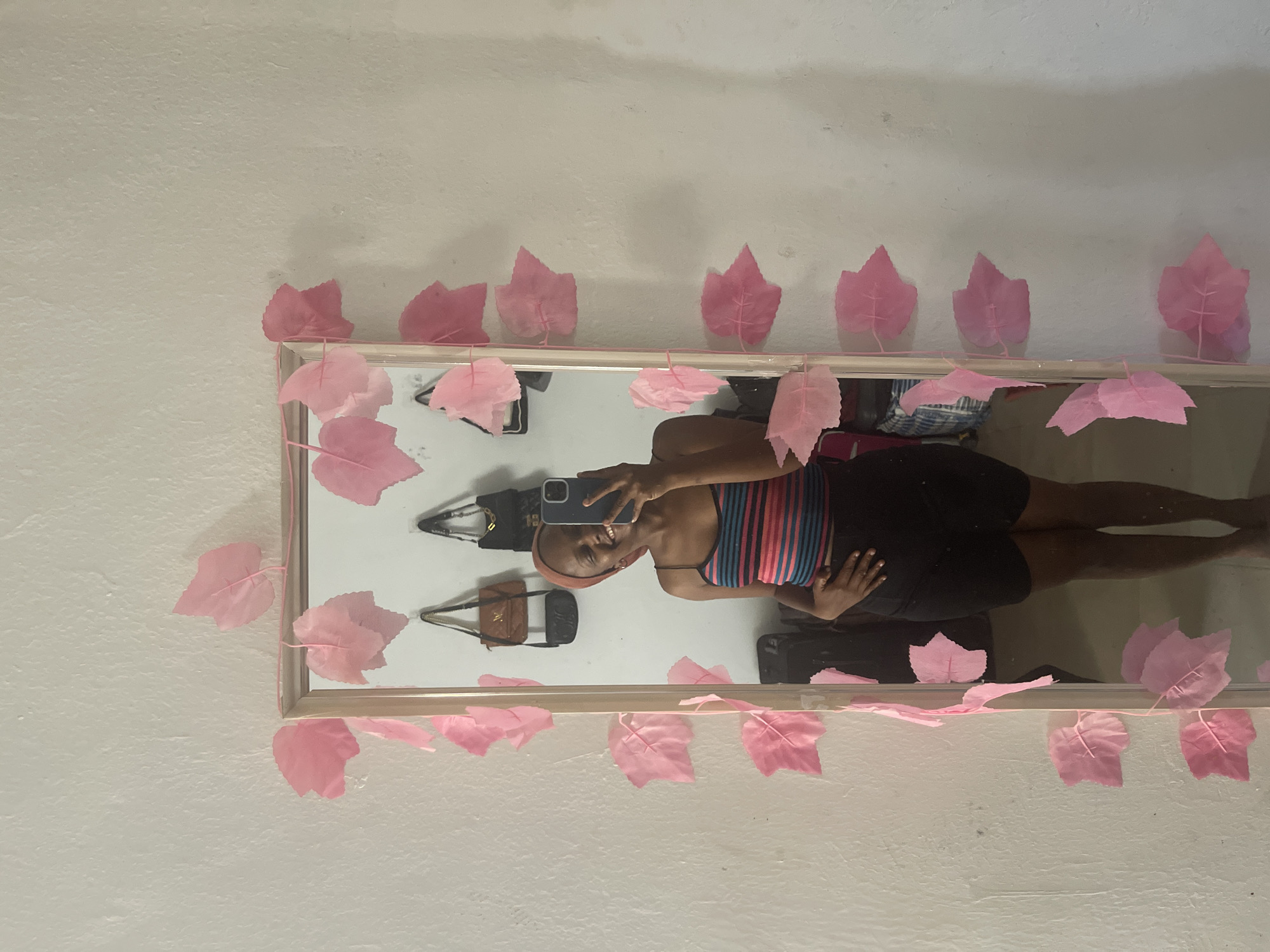 A young woman with dark skin and a headscarf takes a selfie in a full-length mirror. She is wearing a striped tank top with pink, blue, and black stripes and a black skirt. Her left hand is on her hip. The mirror is decorated with a pink leaf garland. In the background, visible in the mirror, are several purses hanging on the wall and some luggage. The wall is white and textured. The scene appears to be indoors, likely in a bedroom, in Jos, Nigeria.