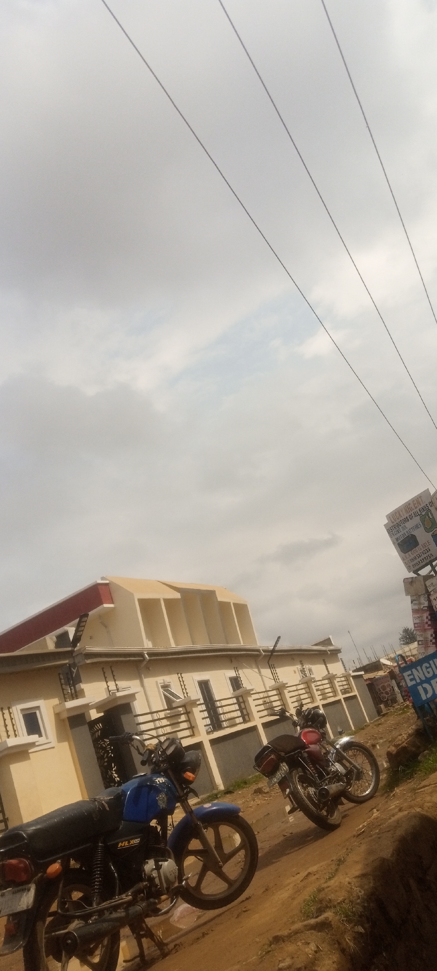 The image depicts a street scene in Sabon Tasha, Nigeria, with two parked motorcycles in the foreground. A blue TVS HLX motorcycle is on the left, and a red motorcycle is on the right. Behind the motorcycles is a two-story building with a beige exterior and a distinctive roof structure. A sign indicating "LUCKY NING ENT DISTRIBUTORS OF ALL KINDS OF ENGINE OIL MOTOR BATTERIES" and contact information is visible on the right side of the image. The sky is overcast, and utility lines are visible in the upper portion of the frame. The ground appears to be a dirt or unpaved surface.