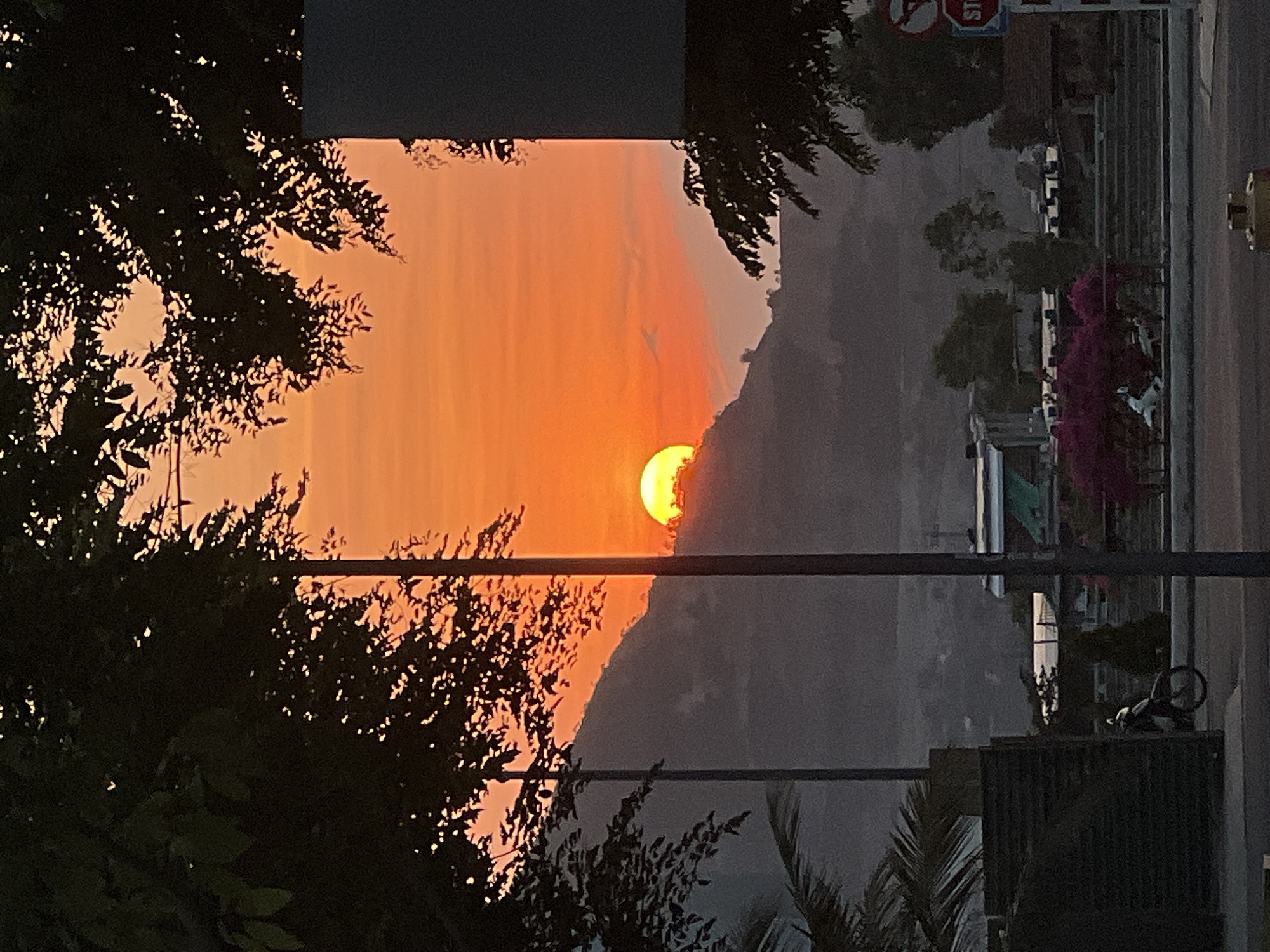 The image depicts a sunset scene in Nam Nha Trang Ward, Vietnam. The sun, a bright orange sphere, is partially obscured by a dark, mountainous silhouette. The sky above the mountain is a gradient of orange hues, suggesting a clear sunset. Trees with dense foliage frame the left side of the image, appearing as dark silhouettes against the bright sky. Utility poles and wires run vertically through the center of the frame, obscuring the view slightly. The right side of the image shows urban elements, including buildings, vegetation, and a road. A "STOP" sign is visible, indicating a street intersection. The overall scene is one of urban development bordering on natural landscapes, with the sunset as the focal point.