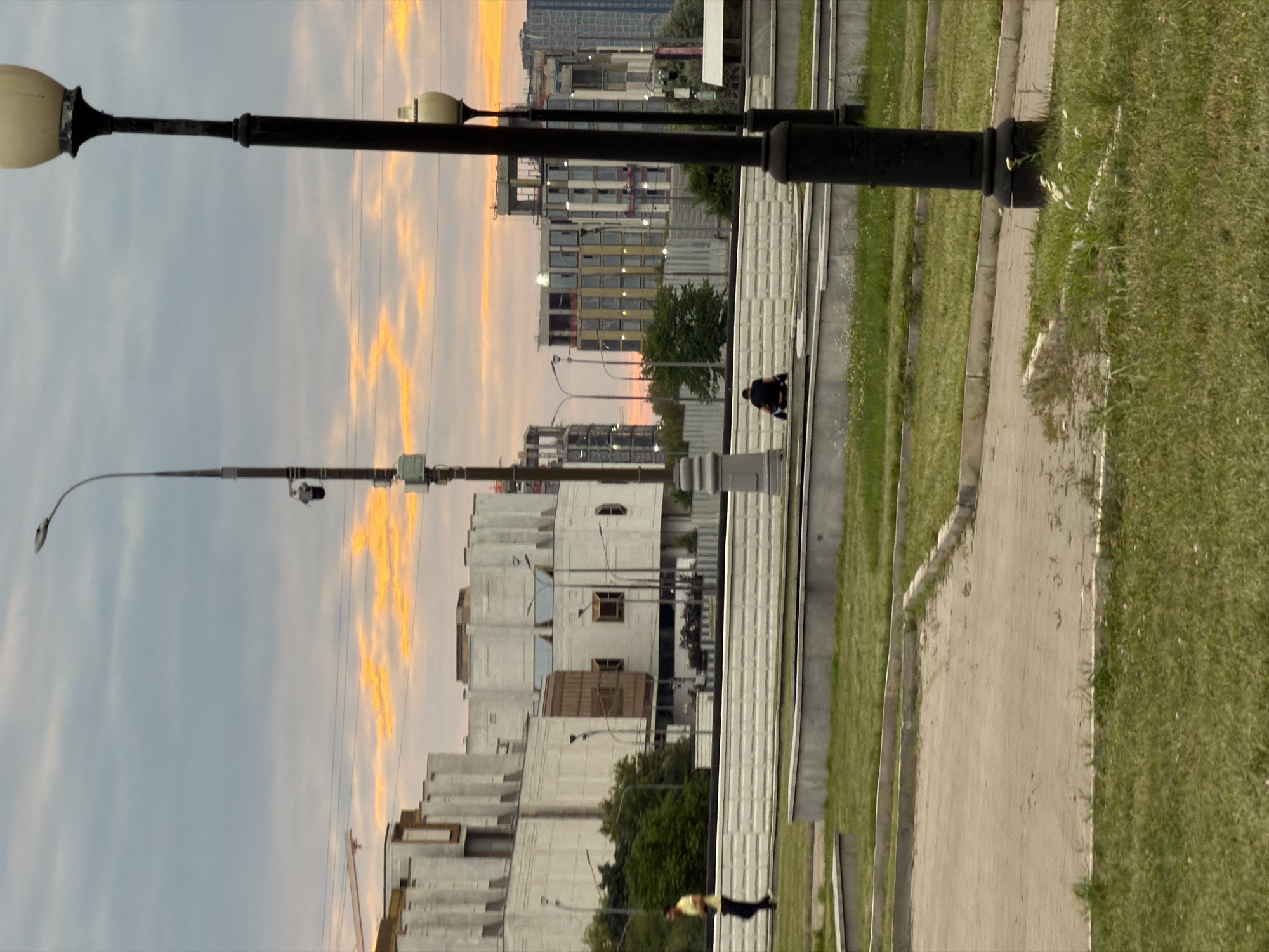 The image depicts a street scene in Moscow, Russia, during a cloudy sunset with streaks of orange light in the sky. In the foreground, a paved area transitions to a grassy lawn with some wildflowers. Two black lampposts, one in the immediate foreground and another further back, flank the scene.
In the mid-ground, a person wearing a yellow shirt and dark pants walks from left to right. Behind them, a white, multi-leveled building complex dominates the background, featuring an industrial-style facade with vertical concrete elements and recessed windows. To the right of this complex, several modern buildings are under construction, showing exposed framework and scattered windows. Another person is seated on a low wall in the mid-ground, facing away from the camera, in front of a concrete barrier. The environment appears to be a park or public space with manicured grass and paved pathways.
