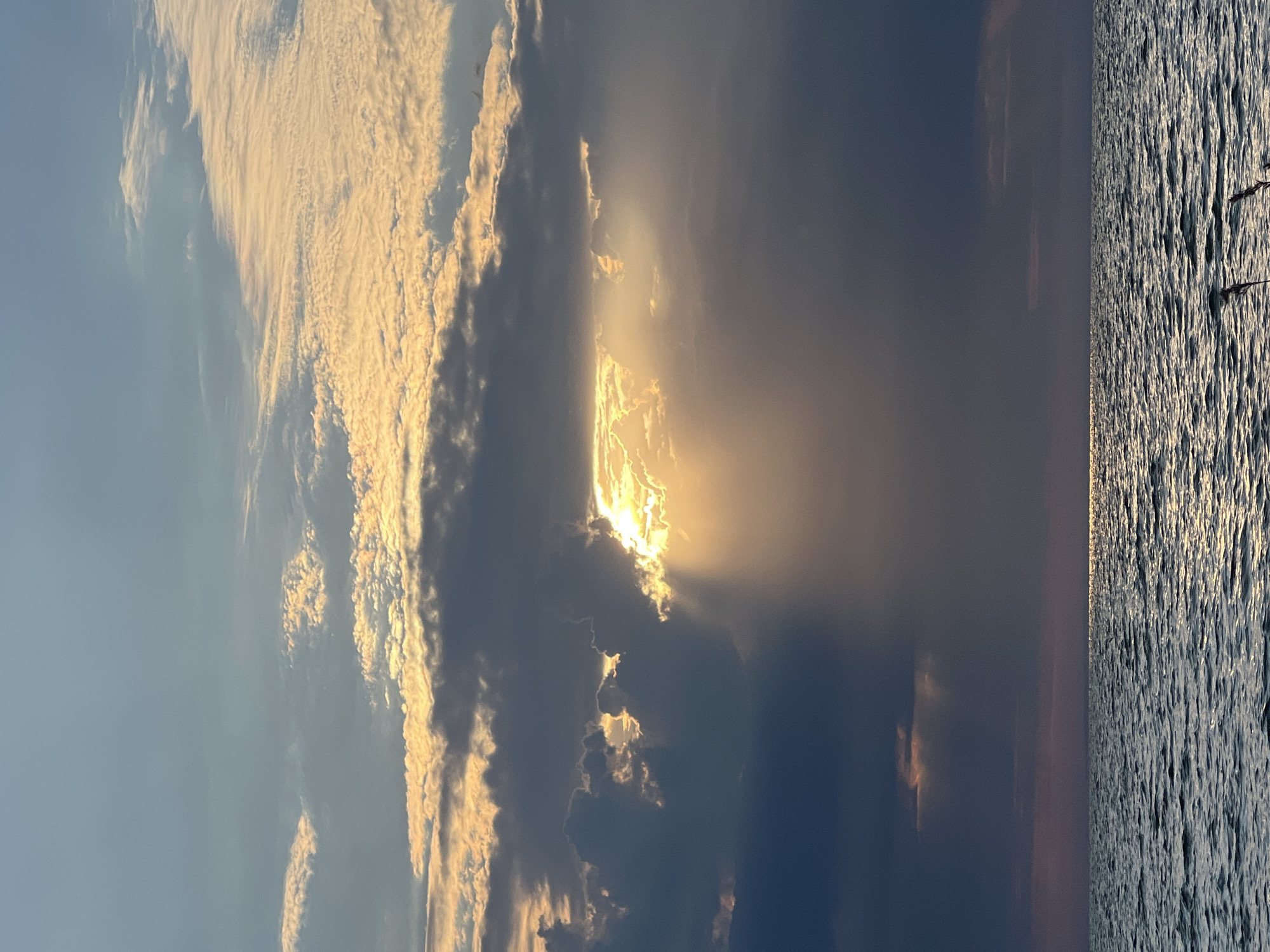 The image depicts a dramatic sunset over a body of water. The sky is filled with a mixture of dark, heavy clouds and lighter, illuminated clouds catching the last rays of the sun. Shafts of sunlight pierce through the clouds, creating a radiant effect. The water below is choppy, reflecting the colors of the sky. There is no indication of the city of Borovoe, Russia, or any people, specific subjects, objects, or actions. The scene is entirely natural and environmental.