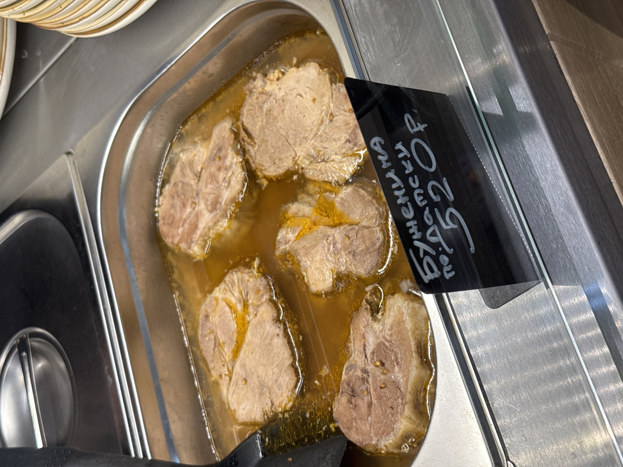 The image shows a close-up of a stainless steel food service container filled with several pieces of cooked meat submerged in a light brown broth. The meat appears to be pork, cut into thick, round slices with some visible fat and seasoning. A black sign with white handwritten text is placed in front of the container, partially obscuring the meat. The text on the sign is in Cyrillic script and reads "Буженина по-Датски 520р." which translates to "Pork roast Danish-style 520 rubles." The background shows the metallic surface of a food counter and stacks of white plates with a beige rim. There are no people visible in the image. The setting appears to be a cafeteria or a restaurant's food display area. The location is not specified to be Yakkima, Russia.