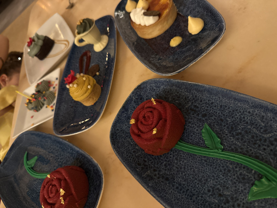 This image captures a dining table laden with multiple plated desserts, consistent with the Be Our Guest restaurant located within Magic Kingdom in Bay Lake, Florida, United States. In the foreground and mid-ground, two dark blue, oblong, textured plates each display a red, rose-shaped dessert with a flocked surface and gold leaf accents, presented on a molded green stem and leaf design. Adjacent, a third dark blue, oblong plate features a yellow, ridged dessert resembling a teacup, topped with a small red flower and white edible pearls, positioned next to a white, tilted, cup-shaped dessert with green frosting and gold flakes. This plate also includes a red sauce drizzle and small white edible spheres. On white, rectangular plates in the background are three gray, spherical cake pops or truffles adorned with multicolored sprinkles, and a dark square cake topped with light green frosting and a red flower. A separate dark blue, square-shaped plate holds a round pastry topped with white whipped cream, nuts, and a dark seed, accompanied by two dollops of yellow cream or sauce. The table surface is uniform light brown. In the out-of-focus background, partial views of two individuals, one wearing a yellow garment, are discernible.