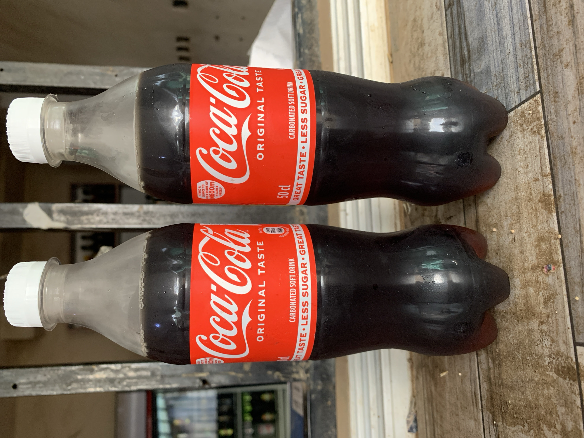 The image shows two bottles of Coca-Cola, each 50 cl in size and labeled as having "Less Sugar." They are positioned side-by-side and slightly overlapping. The bottles have a red label with "Coca-Cola" printed in the classic white font. The background features a somewhat cluttered environment with visible surfaces and what appears to be a refrigerator in the lower portion of the image. Based on the OCR text on the Coca-Cola label, a promotion is visible that relates to winning a share of some prize in cash. It is unclear if this is in the context of the New Stadium, Nigeria.