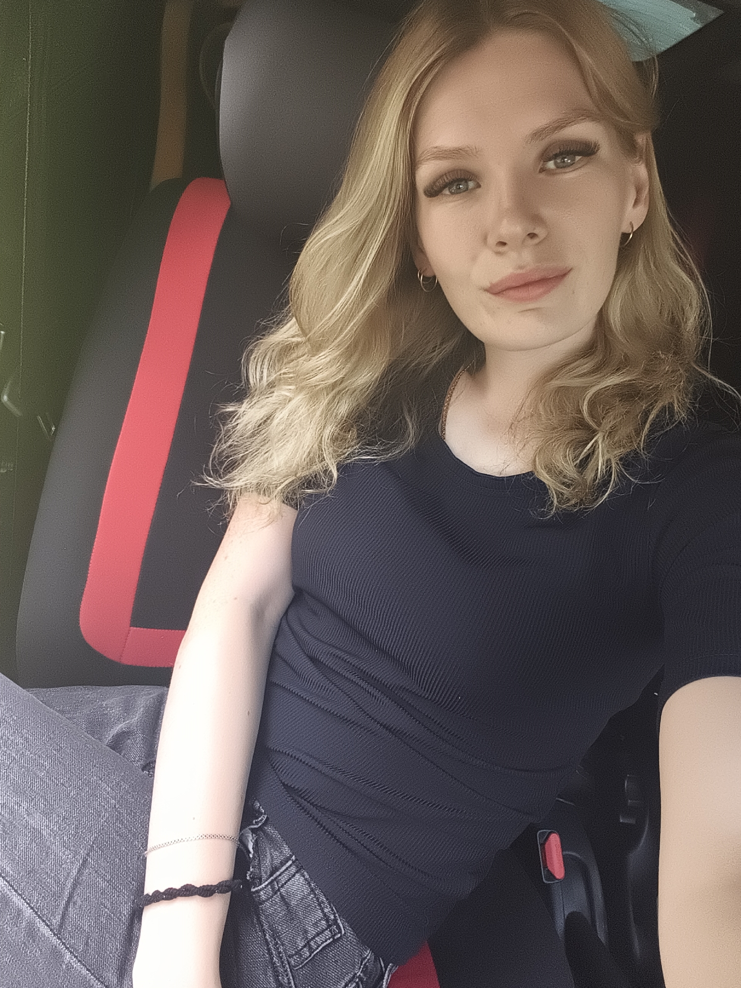 A young woman with blonde, wavy hair is sitting in the driver's seat of a vehicle. She is looking at the camera with a slight smile. She is wearing a black, ribbed, short-sleeved shirt and grey jeans. She has on two bracelets on her left wrist, one silver and one black. The seat next to her has a black cover with a red stripe. The background is dark and out of focus, but appears to be the interior of a vehicle. There is no visible text in the image. The lighting suggests it could be daytime.