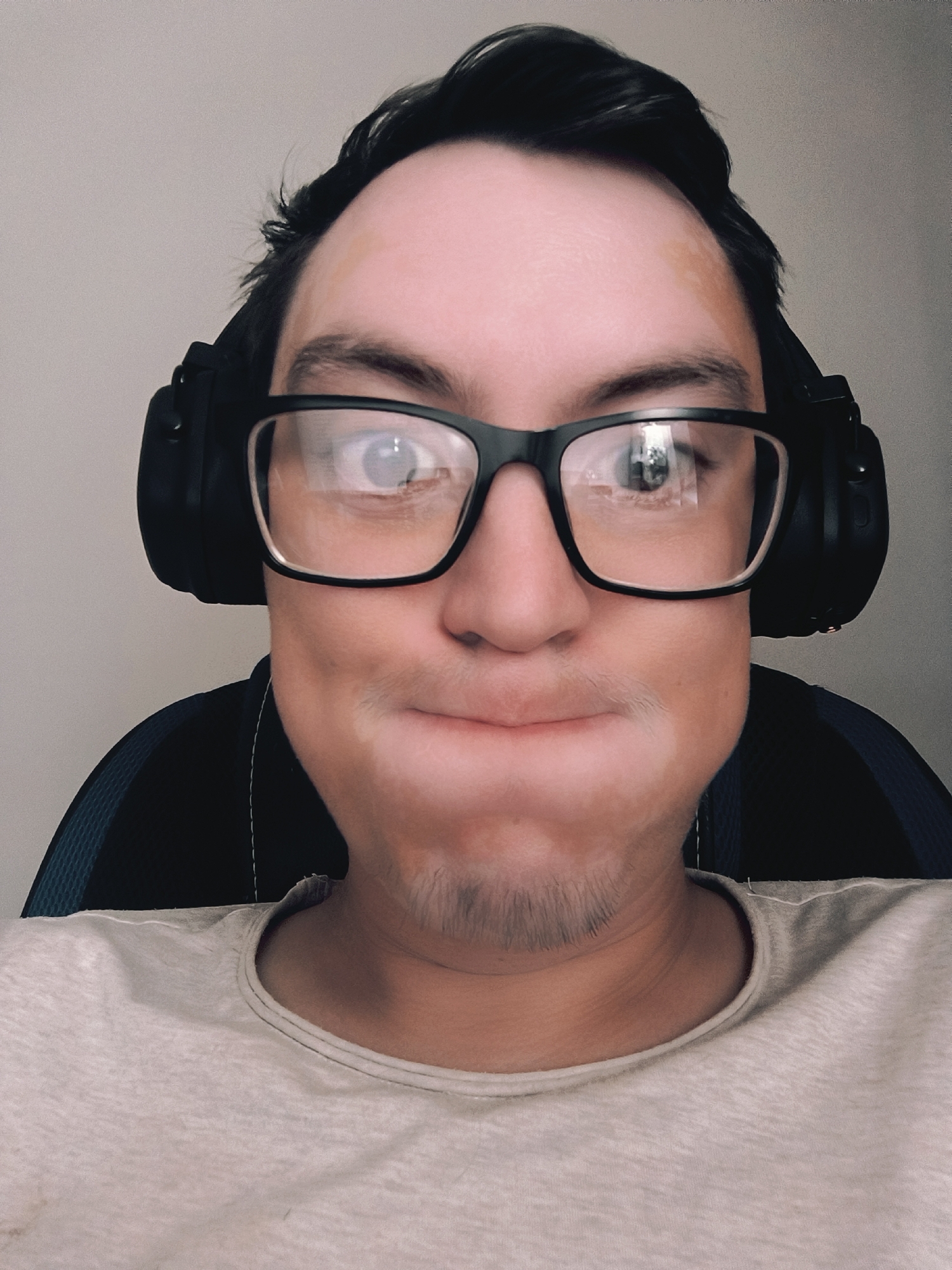 The image is a close-up, head-and-shoulders selfie of a young person wearing black headphones and glasses. The subject has dark hair and a light complexion. Their cheeks are puffed out, and their mouth is pursed. They are wearing a light-colored t-shirt. In the reflection of their glasses, a room with a window is visible. The background is a plain, light-colored wall. The overall impression is informal, possibly taken at home or in a casual setting. There is no visible text in the image.