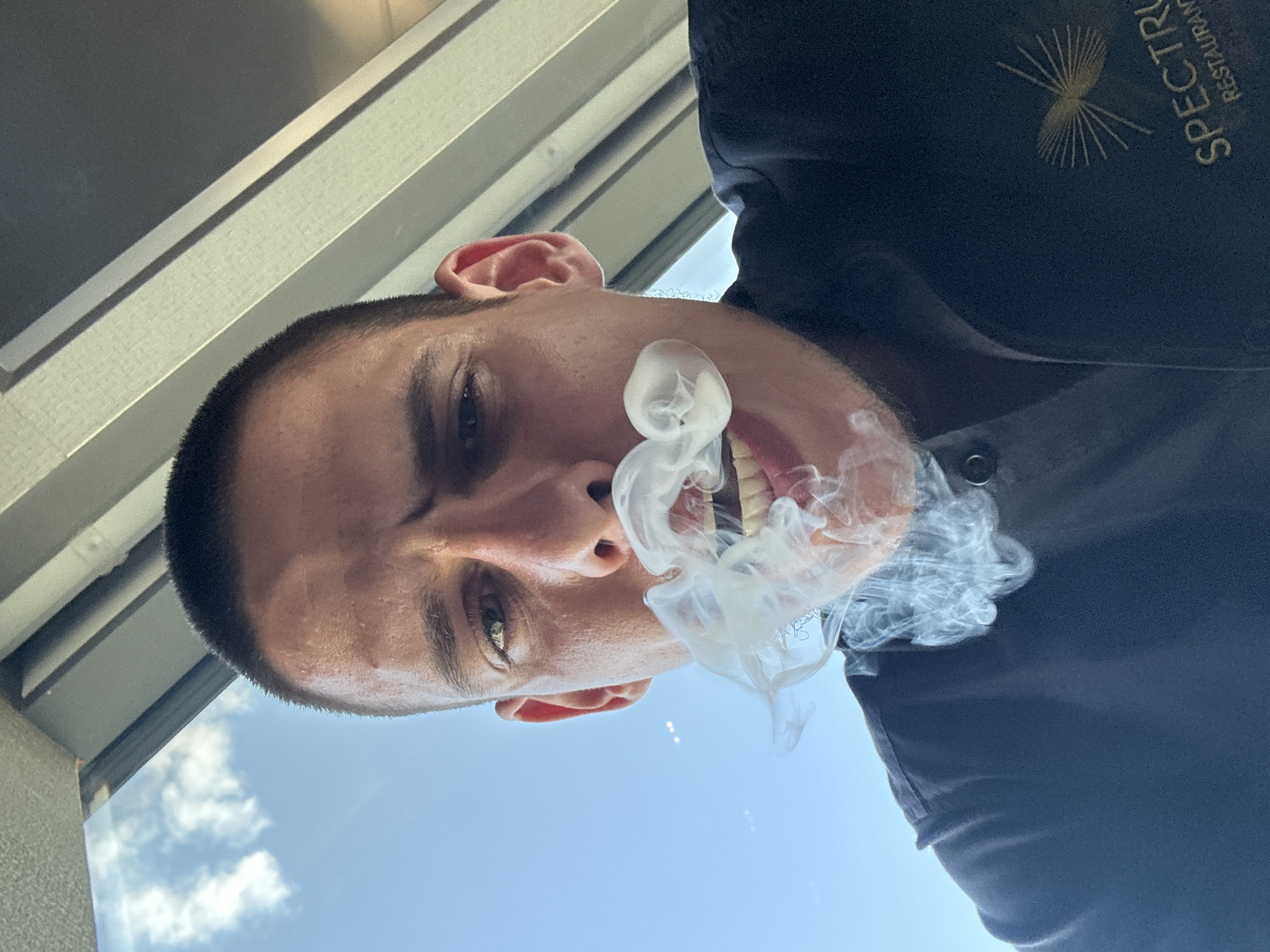 A low-angle, close-up, selfie-style shot features a man exhaling smoke from his mouth. The man has a buzzed haircut and is looking directly at the camera with a slight grimace. He is wearing a dark-colored shirt with a logo that reads "SPECTRUM RESTAURANT". The background shows a bright blue sky with scattered clouds and the angled interior of a building, possibly a restaurant, with light-colored panels and beams. The location in the city of Sofia, Bulgaria is not discernible from the image.