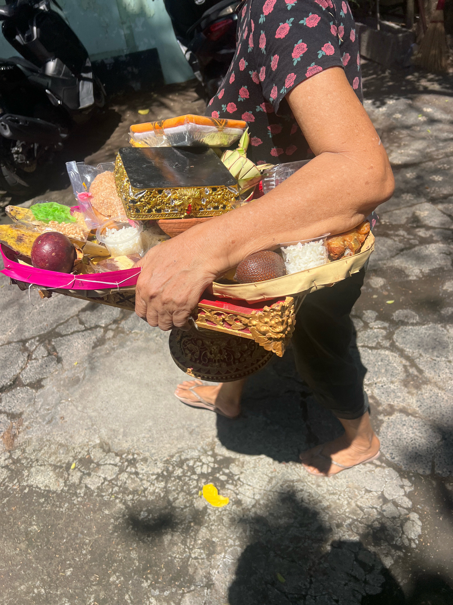In North Kuta, Indonesia, a person walks on a paved outdoor surface, carrying a woven tray filled with offerings. The tray contains a variety of items, including what appears to be rice, fruit, crackers, and other food items, some of which are enclosed in plastic packaging. A decorative, gold-colored box is positioned in the center of the tray. The person is wearing a black t-shirt with a floral pattern and dark pants, and is barefoot, wearing simple flip-flops. In the background, motorcycles are visible, suggesting an urban or semi-urban setting. The ground is a mix of concrete and asphalt, with some areas showing wear and tear. Sunlight casts shadows on the ground, indicating it is daytime. A single yellow petal is visible on the ground near the person's feet.