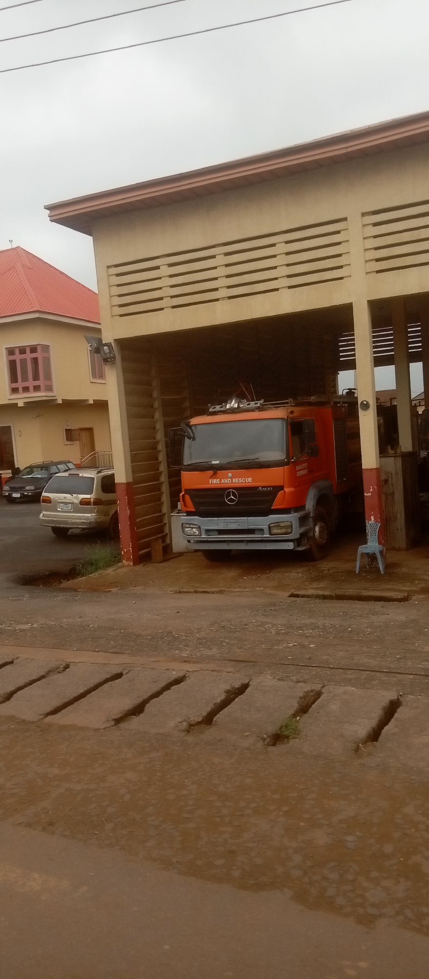 The image depicts the Enugu State Fire Service building in Enugu, Nigeria. An orange fire truck is parked under an open-sided structure, which appears to be a bay for housing vehicles. The truck has "FIRE AND RESCUE" written on its front. To the left of the fire truck, a yellow building with a red roof is visible, likely part of the same facility. Several cars are parked in front of the yellow building. The ground in the foreground is a paved road with drainage channels. The sky is overcast.