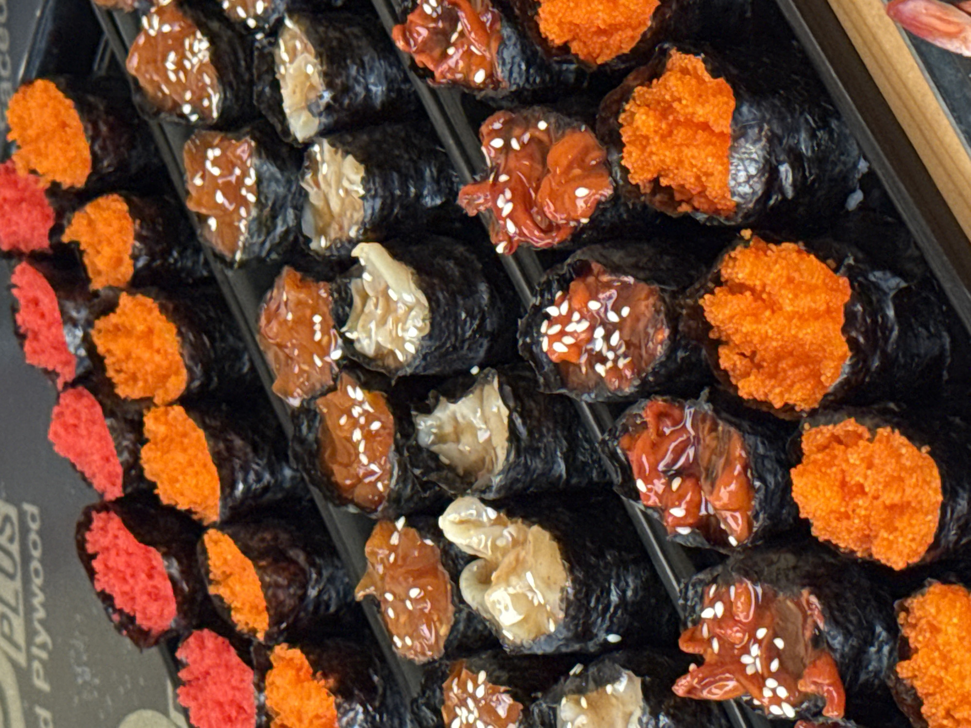 A close-up, top-down, angled view shows an assortment of sushi rolls arranged in rows on a black tray. The sushi rolls are small, cylindrical, and wrapped in dark seaweed. The toppings vary in color and texture, including bright red and orange roe, a glistening brown sauce with sesame seeds, and what appears to be shredded white seafood or vegetable. The lighting is bright and even, highlighting the vibrant colors of the sushi. The background includes a dark surface with some text that is partially visible, reading "d Plywood". The sushi appears to be displayed for sale, likely at a market or food stall. The scene is clean and the food is presented attractively. The overall impression is of fresh, ready-to-eat sushi.