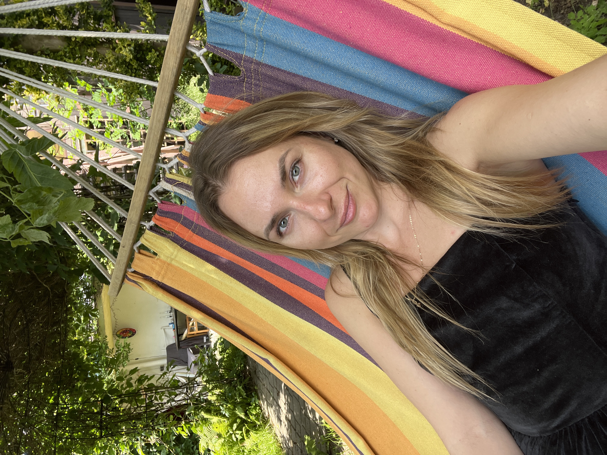 A blonde woman with green eyes and fair skin is reclining in a colorful striped hammock outdoors. She is wearing a black sleeveless top and a delicate gold necklace. Her hair is long and blonde, falling around her shoulders. The hammock is suspended between two wooden poles and is made of a fabric with horizontal stripes of orange, yellow, purple, blue, and pink. In the background, lush green foliage and trees suggest a garden setting. Partially visible in the background is a building with white walls and a colorful circular decoration on it. To the left, a paved pathway is visible, shaded by overhead leaves. The scene appears to be set in a relaxed, outdoor environment, likely a backyard or garden, possibly in Krakow, Poland, given the presence of what appears to be a Polish building in the background. The woman is looking towards the camera with a slight smile.