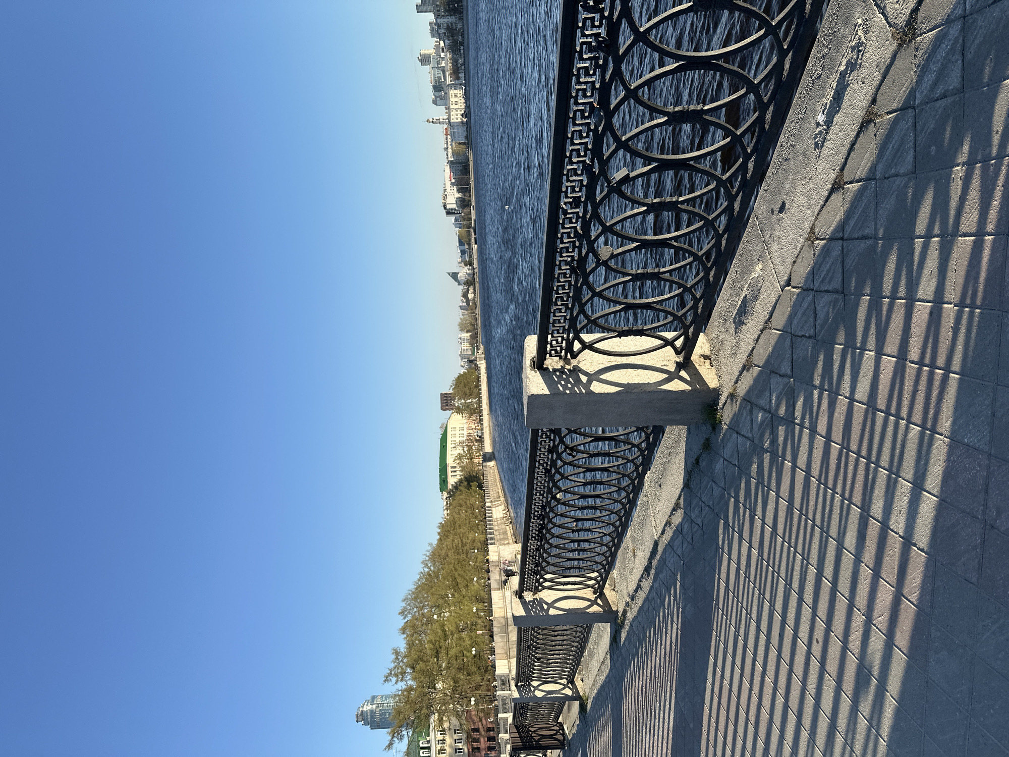 This image presents a scenic view of a river embankment in Yekaterinburg, Russia, captured on a clear, sunny day. The scene is dominated by a wide, dark blue river on the left side, its surface gently rippling. Across the river, the distant bank is lined with various buildings, ranging from multi-story structures to what appear to be taller, more modern edifices, along with a continuous stretch of green trees.
To the right, a broad pedestrian walkway or promenade extends into the distance. It is paved with square or rectangular grey tiles, onto which long, defined shadows are cast by an ornate, dark-colored metal railing that runs along the river's edge. This decorative railing, likely made of cast iron, features a pattern of interlocking circles and other geometric designs, acting as a barrier between the walkway and the water. Several dark benches are placed along the promenade, behind the railing.
The sky above is a brilliant, cloudless blue, transitioning to a lighter hue towards the horizon. The distinct, elongated shadows suggest either early morning or late afternoon. A few small, indistinct figures can be seen in the distance along the embankment, indicating people are enjoying a stroll or resting. The overall impression is one of peaceful urban beauty under pleasant weather conditions. No visible text is present in the image.