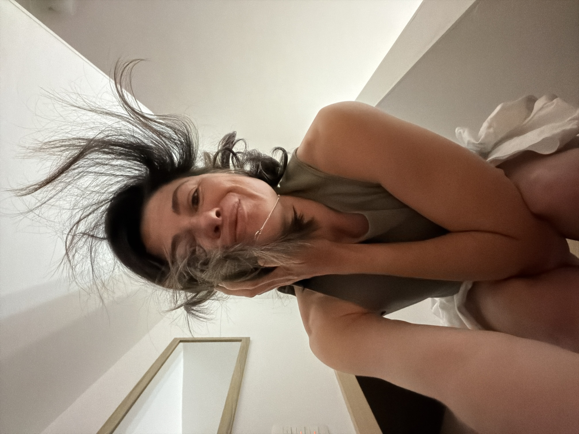 The image is taken from a low angle, looking up at a woman with dark hair. Some of her hair is styled upwards. She is wearing a sleeveless top. Her arms are raised towards her face. She is smirking. A gold chain is visible around her neck. In the background, there is a white wall and a framed object. No information is available to determine this image was taken in Budva, Montenegro.
