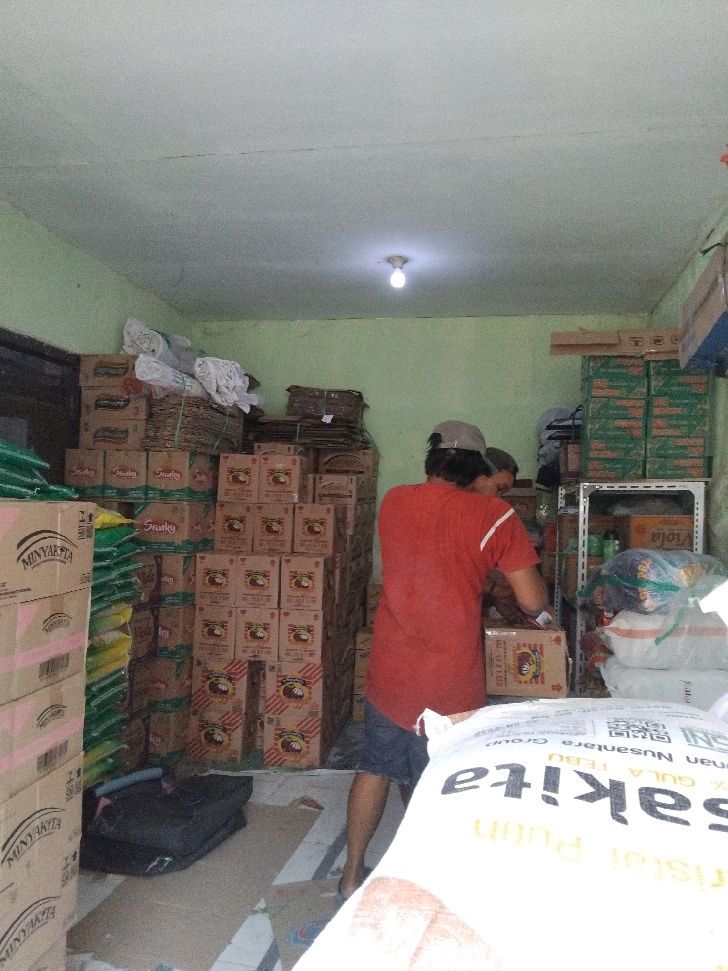The image depicts the interior of a small store or warehouse located in Berbek, Indonesia, likely a retail outlet or a local distribution point. The room is filled with stacks of boxed goods, primarily food products like cooking oil and sugar, indicated by labels such as "Minyakita" and "Gula Kristal Putih." The packaging suggests these are consumer-sized products.
In the foreground, a large white sack with Indonesian text, "Gula Nusantara Sakita", is prominently displayed, partially obscuring a section of the room. The text translates to "Nusantara Sakita Sugar." The sack is likely full of sugar.
Two individuals are present in the scene. One person, wearing a red t-shirt and dark shorts, is seen from the back and side, appearing to be handling or organizing boxes. They are wearing a cap. Another individual is partially visible behind them, suggesting collaboration or shared work in the store.
The environment appears functional rather than elaborate. The walls are painted a light green, and the ceiling is textured white with a visible light fixture. Cardboard boxes are stacked high, creating narrow aisles. Some boxes are neatly arranged, while others appear to be in disarray, suggesting ongoing activity or storage.
A metal shelving unit is visible on the right side of the image, holding more boxed goods and bags. On the floor, a dark-colored folded item, possibly a bag or mat, is present, along with loose cardboard pieces. The overall impression is that of a busy, operational retail or storage space dealing with consumer goods. The lighting is ambient, with the overhead light providing illumination. The presence of "Minyakita" and other Indonesian brand names clearly places the scene within Indonesia.