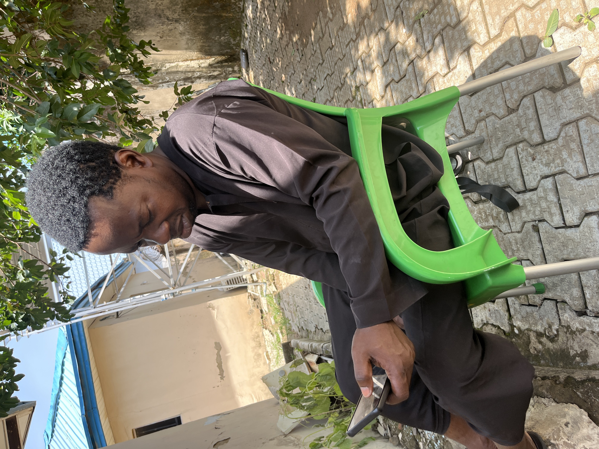 An outdoor medium shot captures a man seated on a bright green plastic chair, engrossed in his smartphone. He wears a dark brown robe with wide sleeves and loose-fitting trousers. The man's head is slightly tilted down, his gaze fixed on the device held in his left hand. His right hand rests on his lap. He is positioned outdoors, with a background featuring a light beige building with a blue-tiled roof and a concrete wall. Sunlight casts dappled shadows across the textured, light-grey paving stones that make up the ground. A cluster of green leaves from an overhanging tree provides shade. The overall scene suggests a relaxed setting, possibly a courtyard or garden area within Abuja, Nigeria.