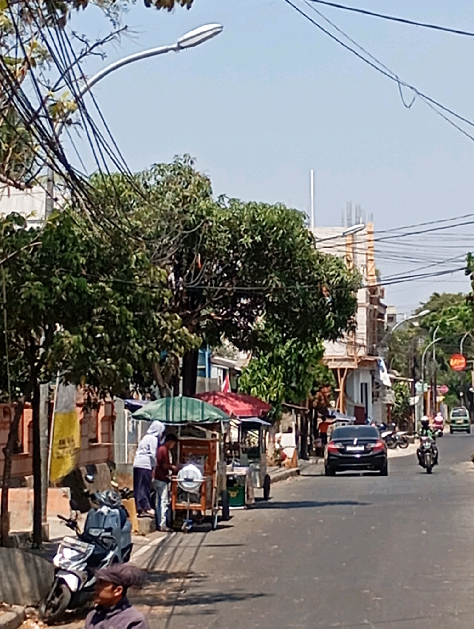 In Cimahi, Indonesia, on a sunny day, a street scene unfolds. A dark sedan is driving down the road, followed by a motorcycle. On the side of the road, a food cart is set up, with two people attending to it. A scooter is parked nearby, and a man wearing a cap is looking forward. In the background, a building is under construction, with scaffolding visible. The street is lined with trees and utility poles, with a street lamp arching over the road.