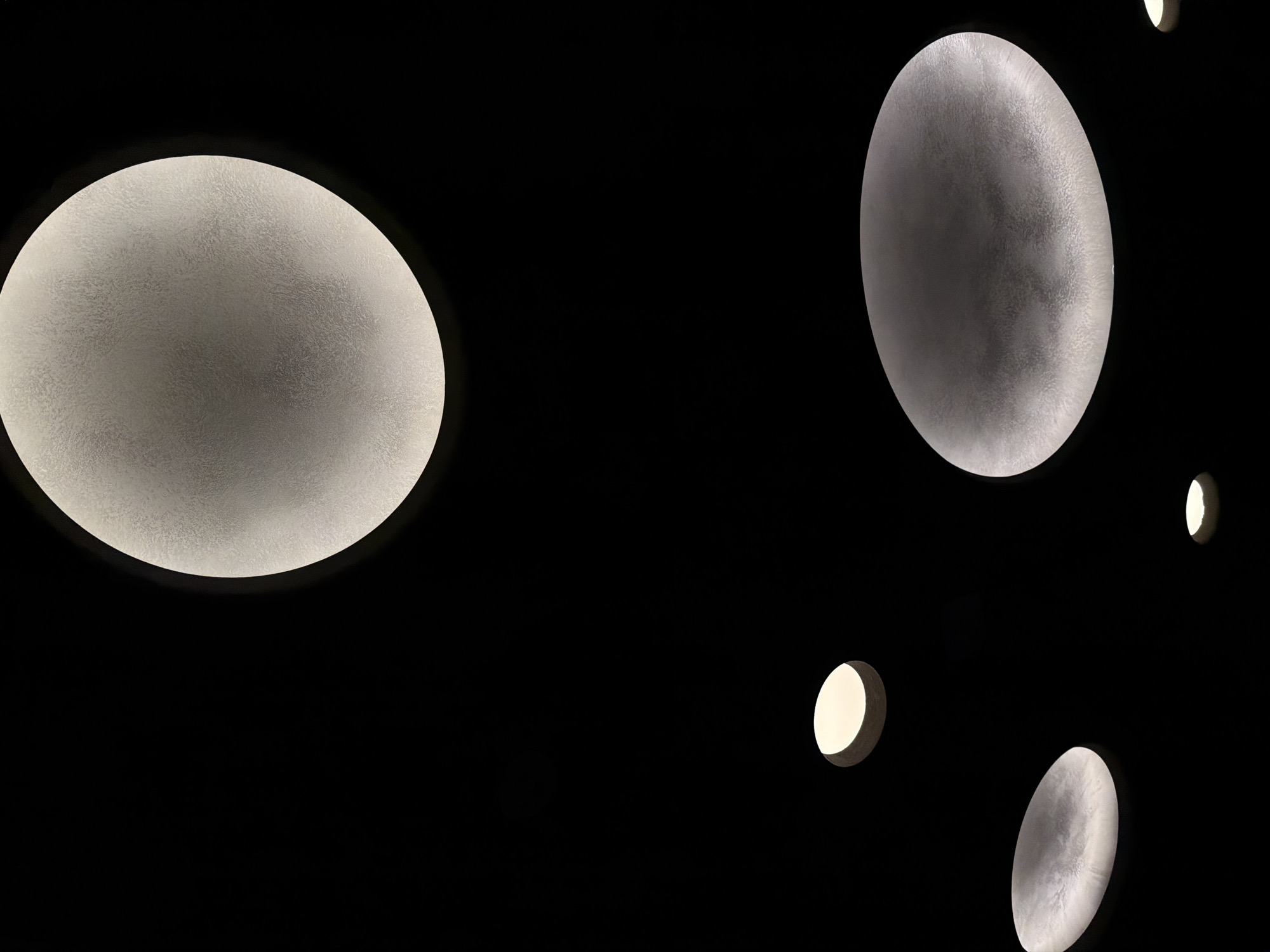 The image shows a dark ceiling with several round, diffuse light fixtures. A large, nearly circular light fixture dominates the left side of the frame. Another elongated oval light fixture is positioned on the right. Two smaller, circular light fixtures are also visible. The background is completely black. The image may be taken in Izhevsk, Russia, but there is no information about the city itself evident in the scene.