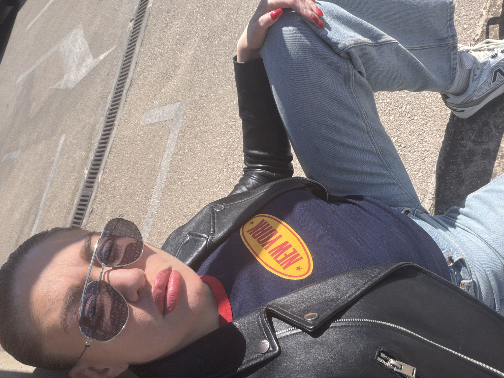A person is lying on a paved surface outdoors, wearing blue jeans, black boots, a dark blue shirt with a "NEW YORK" graphic, and a black leather jacket. They are also wearing sunglasses. The pavement has painted arrows and a drainage grate. The location is the city of Bar, Montenegro.