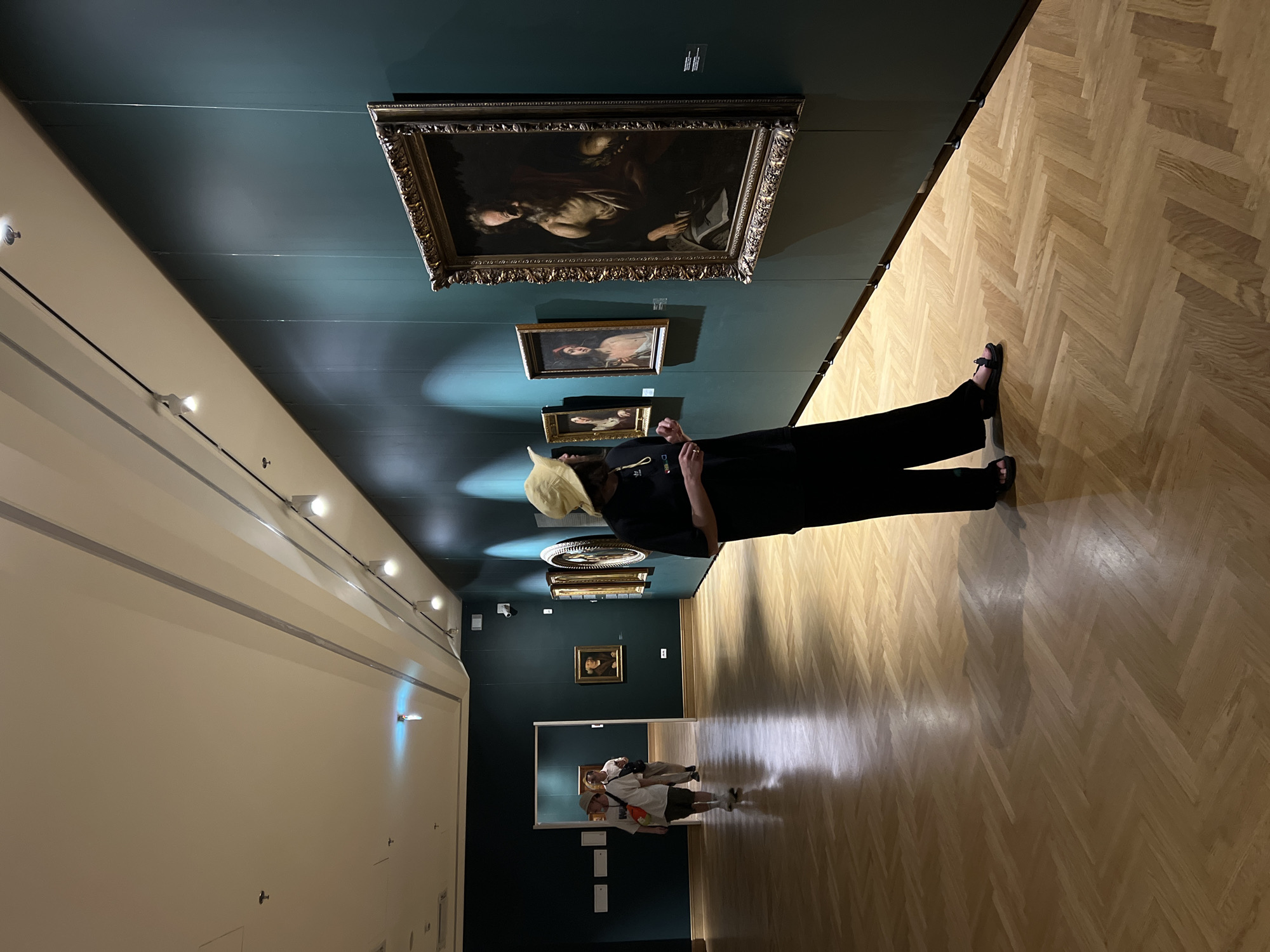 The image depicts the interior of a building, potentially a museum, with a dark green wall displaying several framed paintings. A person wearing a tan hat, a black shirt, and black pants is standing in the foreground, facing the paintings. The floor is a light-colored herringbone parquet. Further down the hall, there are two other people near what appears to be a mirror. The wall opposite the paintings is a light color with several light fixtures. Based on context clues like the artwork, and parquet style, the photo may have been taken at the National Museum in Belgrade, Serbia.