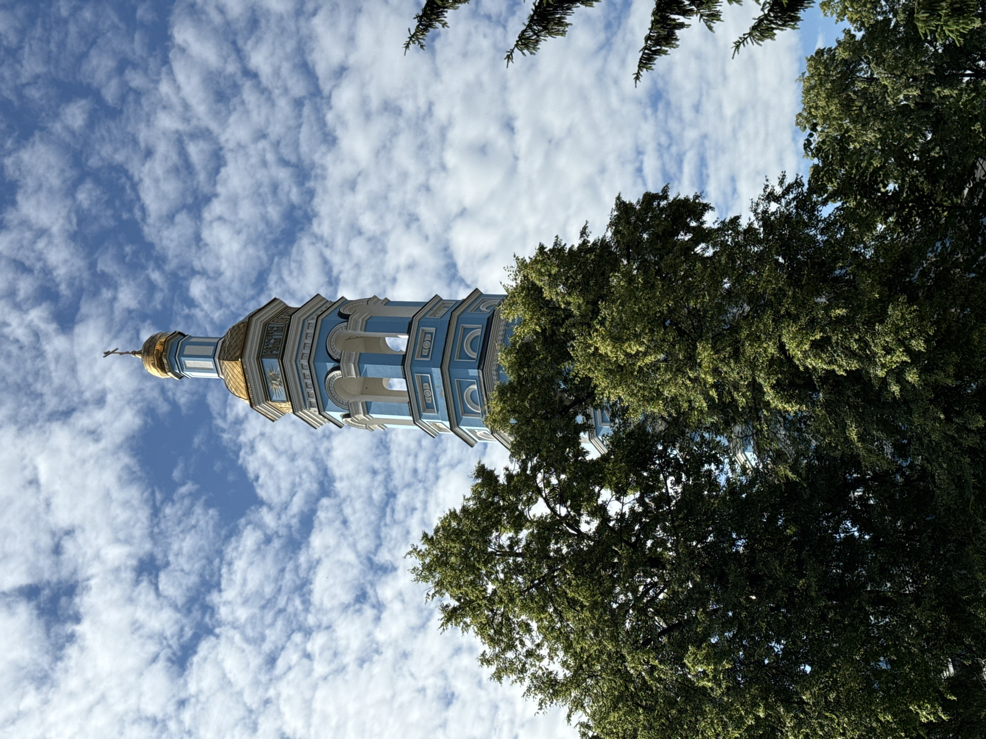 An upward-facing shot captures a tall, slender bell tower with blue and white accents and a gold dome topped with a cross. The tower stands against a bright blue sky dotted with fluffy white clouds. Lush green trees flank the lower portion of the tower, their branches reaching upwards. The overall scene depicts a serene and picturesque view of a church bell tower amidst nature.